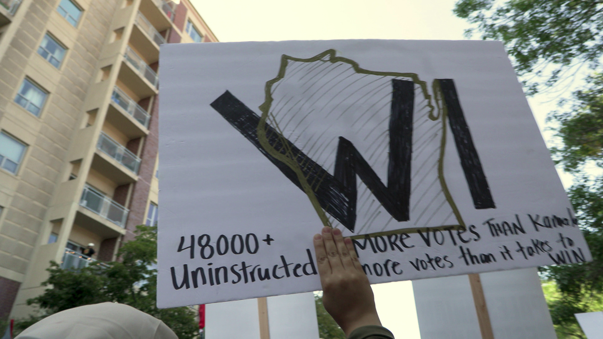 A hand holds a sign with an outline of Wisconsin and the words "48000+ uninstructed" and "more votes than Kamala, more votes than it takes to win" with a building and trees in the background.