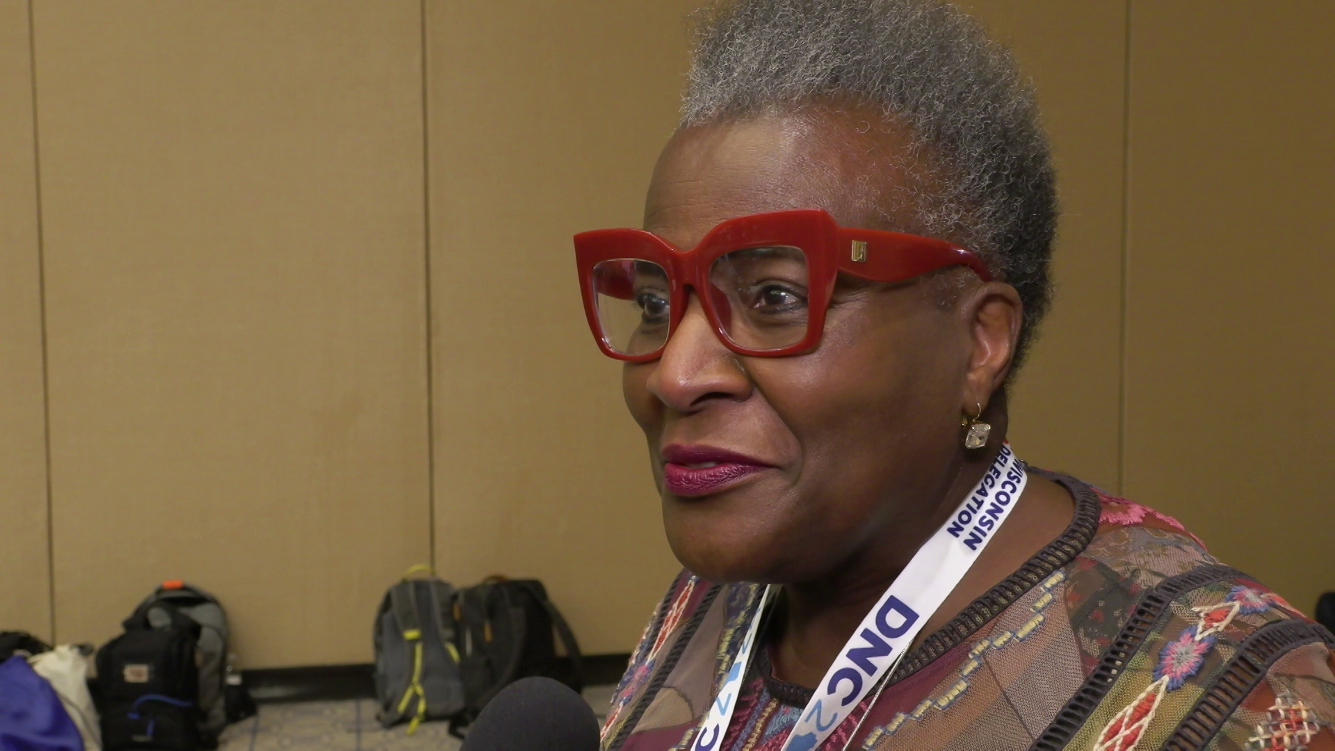 Thelma Sias stands and speaks indoors with backpacks on the floor in the background.