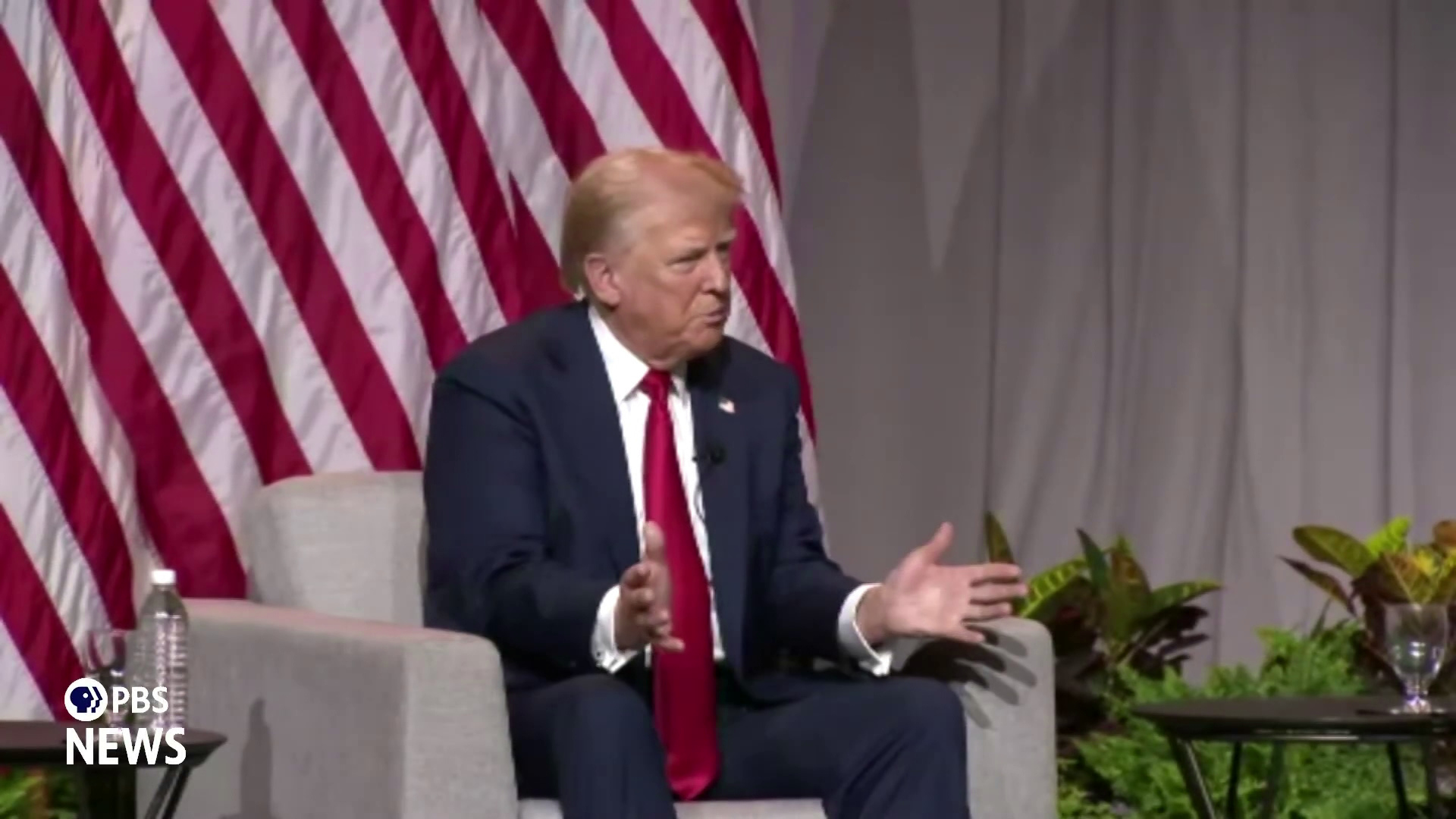 Donald Trump gestures with both hands and speaks while sitting in front of a U.S. flag and potted plants and next to a table with a bottle.