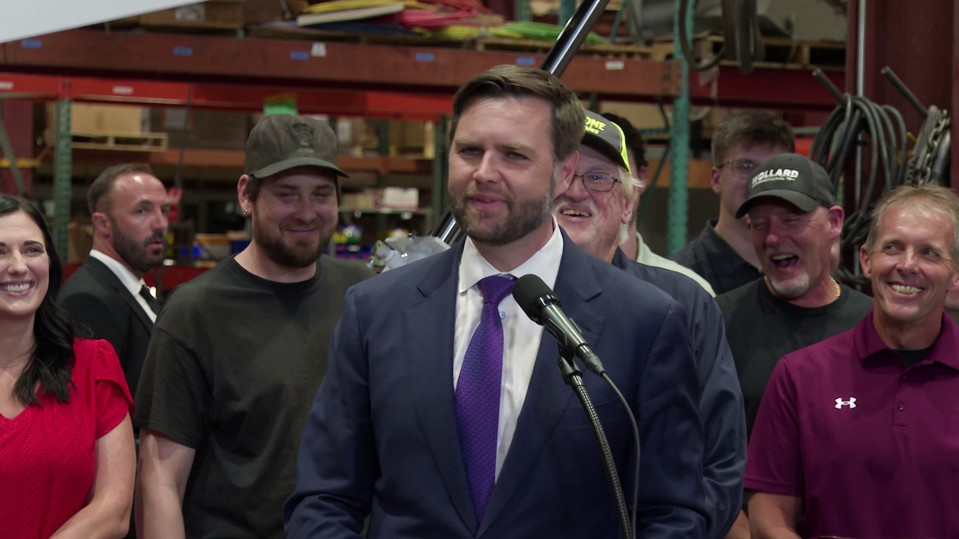 JD Vance stands and speaks behind a podium with a microphone in front of a group of people standing behind him with metal beams cables in the background.