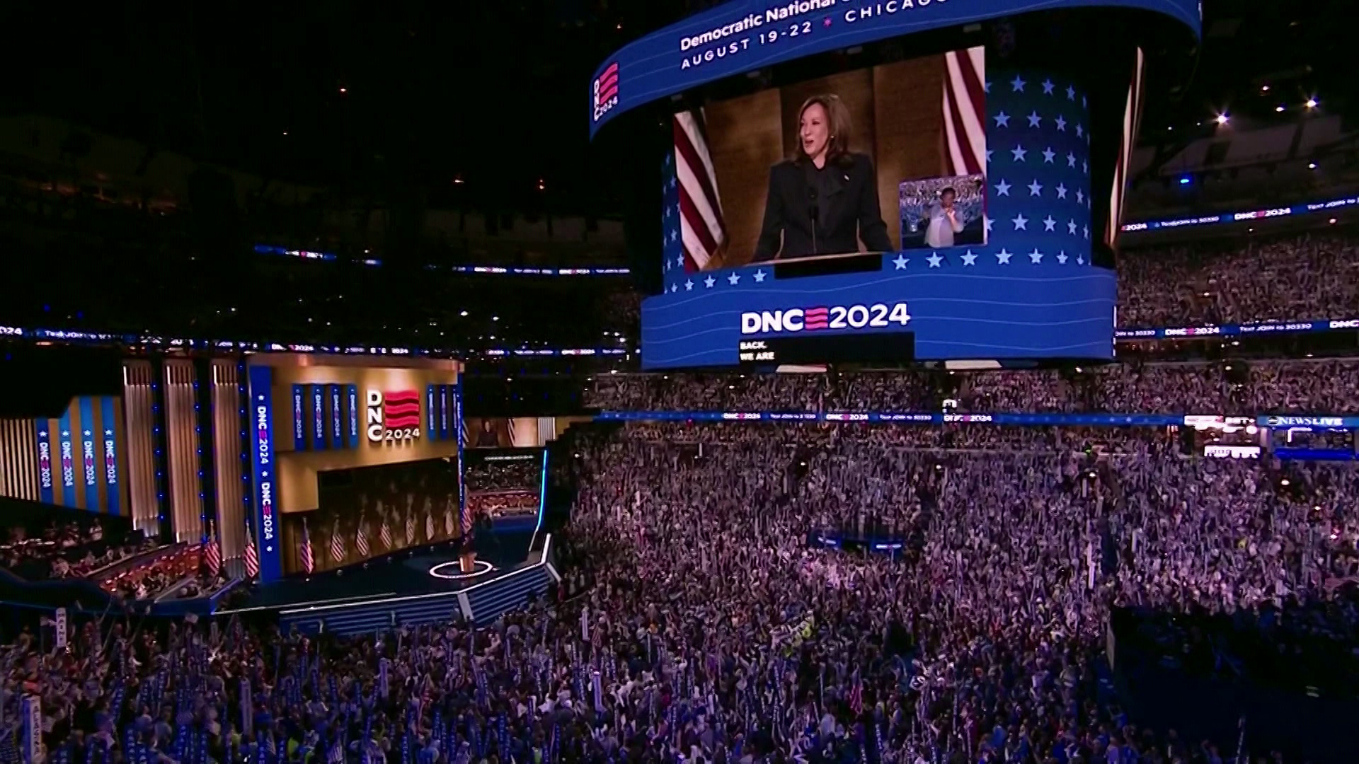 A muti-sided electronic stadium display with a screen showing Kamala Harris speaking and a "DNC 2024" logo is mounted to the ceiling of an arena filled with thousands of people facing a stage at one end with Harris standing behind a podium.