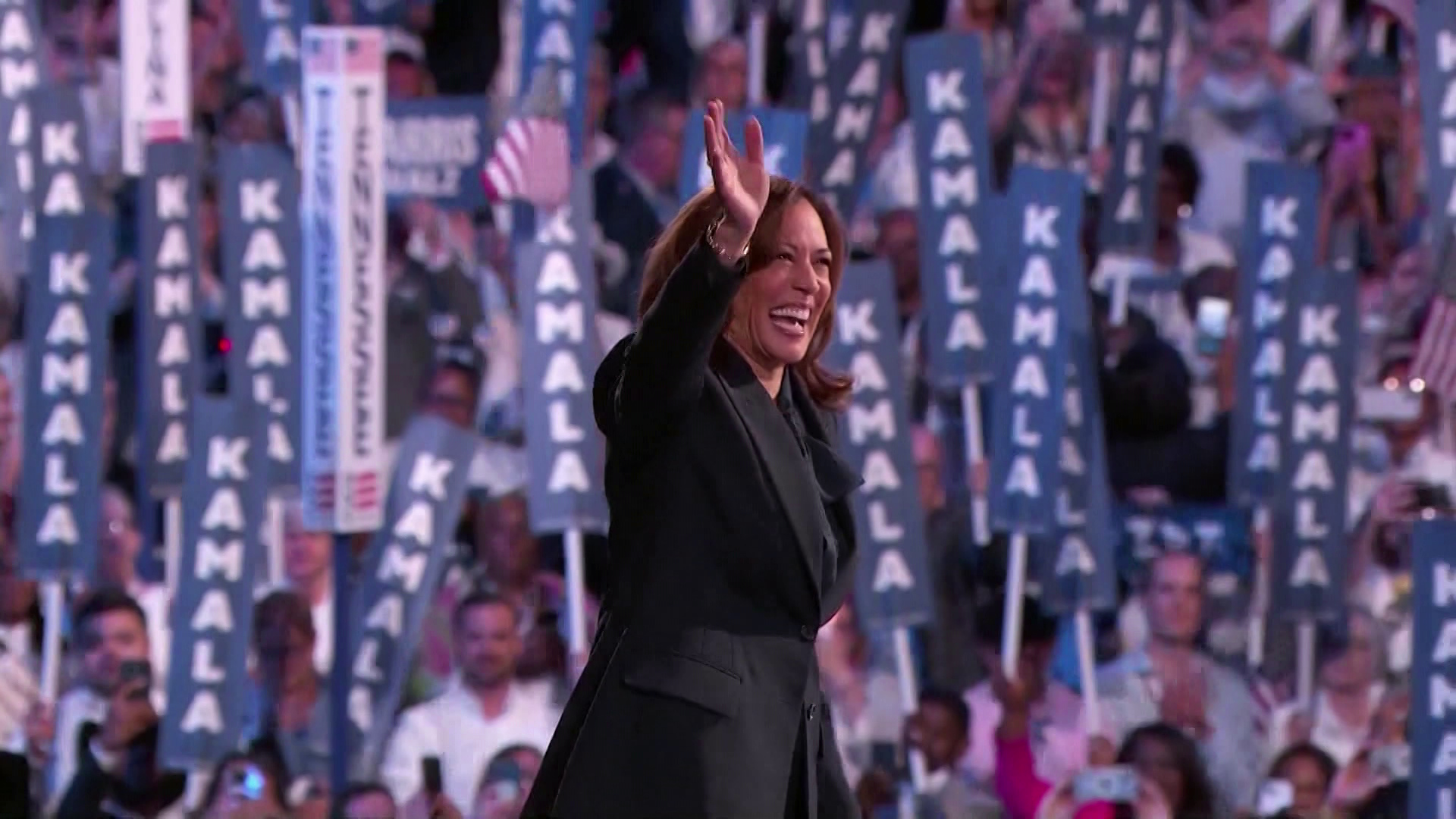 Kamala Harris stands and waves with an out of focus crowd of people holding up signs reading "Kamala" in the background.