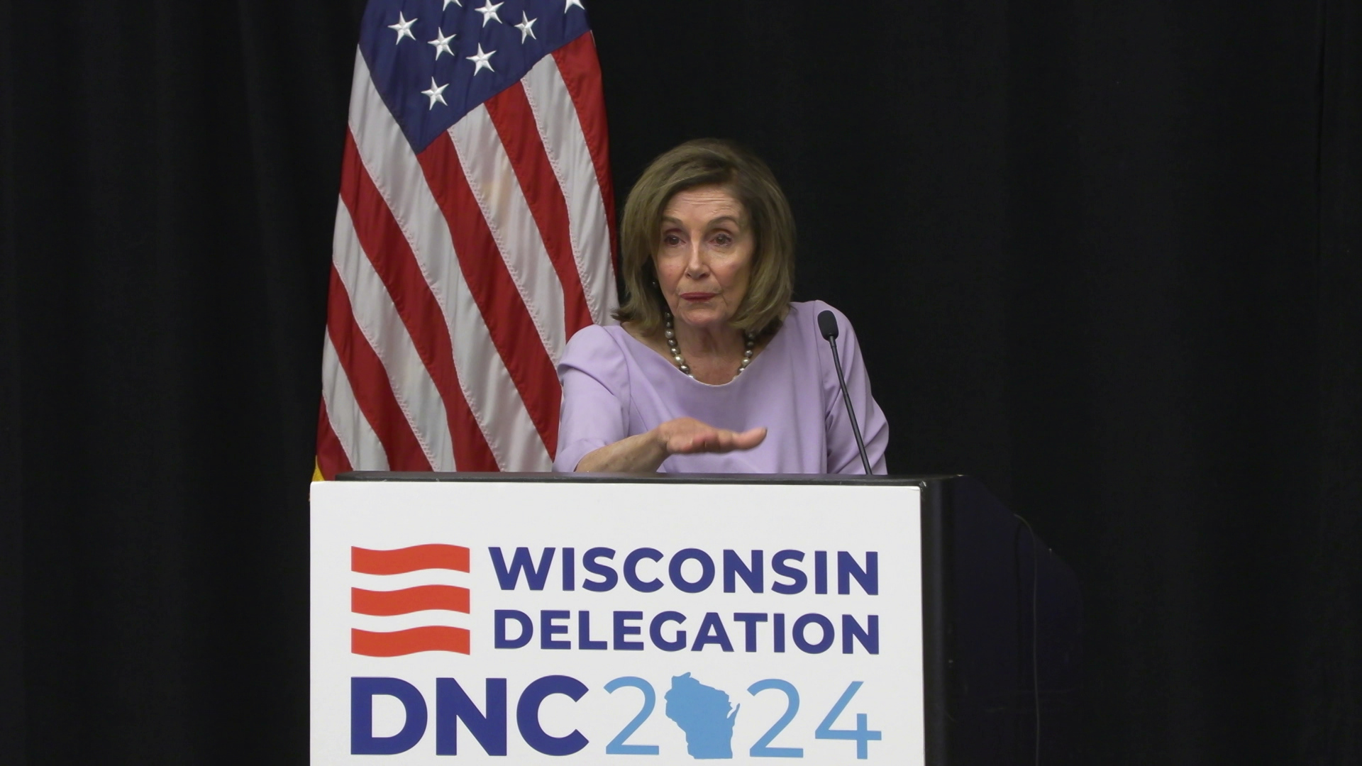 Nancy Pelosi gestures with her right hand and speaks into a microphone while standing behind podium with a sign on the front that reads "Wisconsin Delegation DNC 2024," with a U.S. flag in the background.