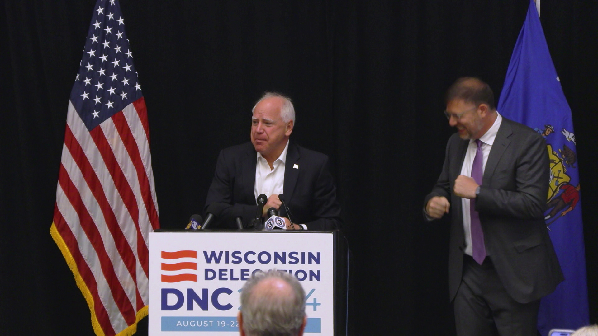 Tim Walz speaks multiple microphones on the surface of a podium with a sign reading "Wisconsin Delegation Breakfast DNC 2024" next to Ben Wikler dancing, with the U.S. and Wisconsin flags in the background.