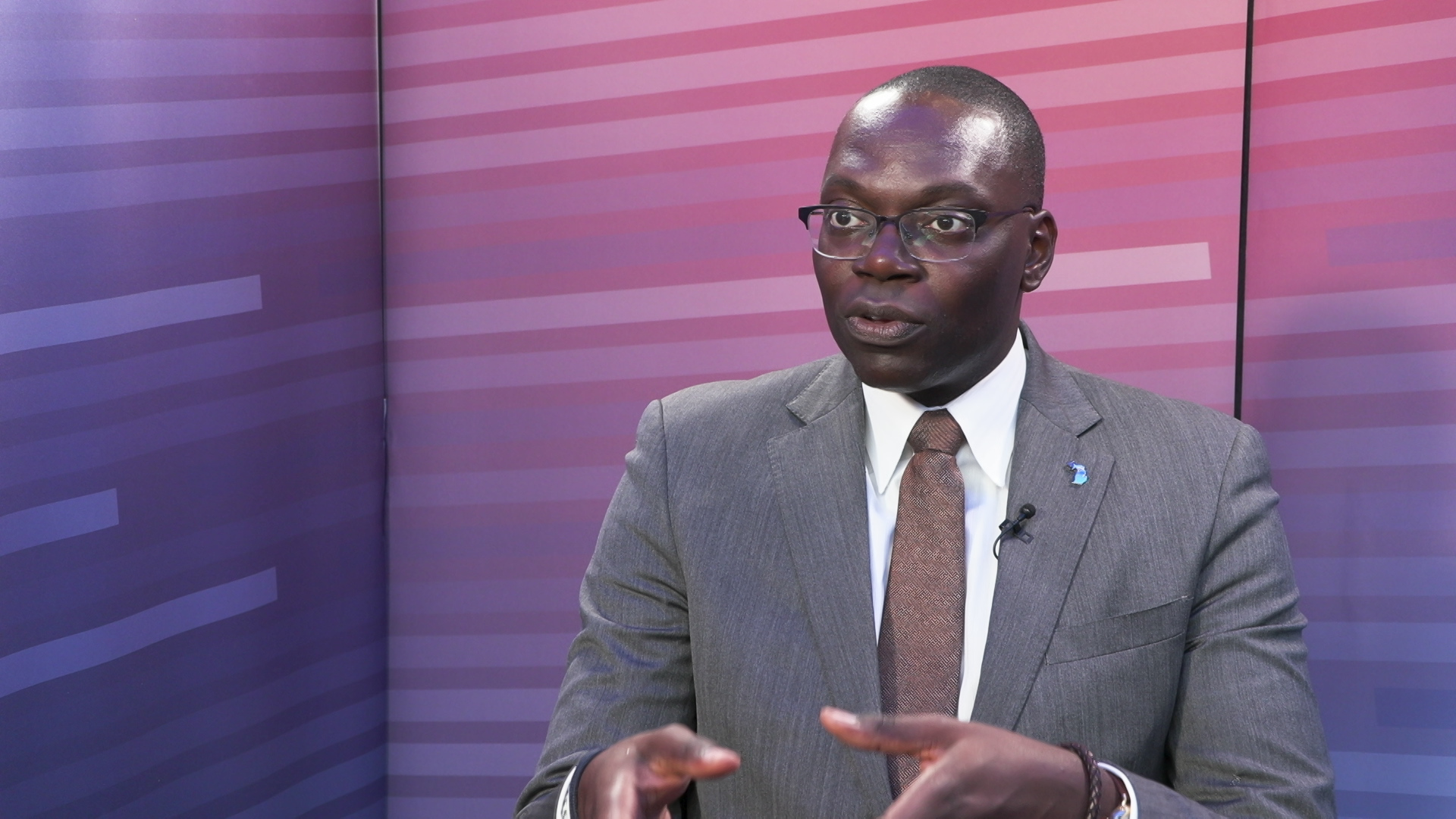 Garlin Gilchrist speaks while sitting and gesturing with hands in front of a gradient background.