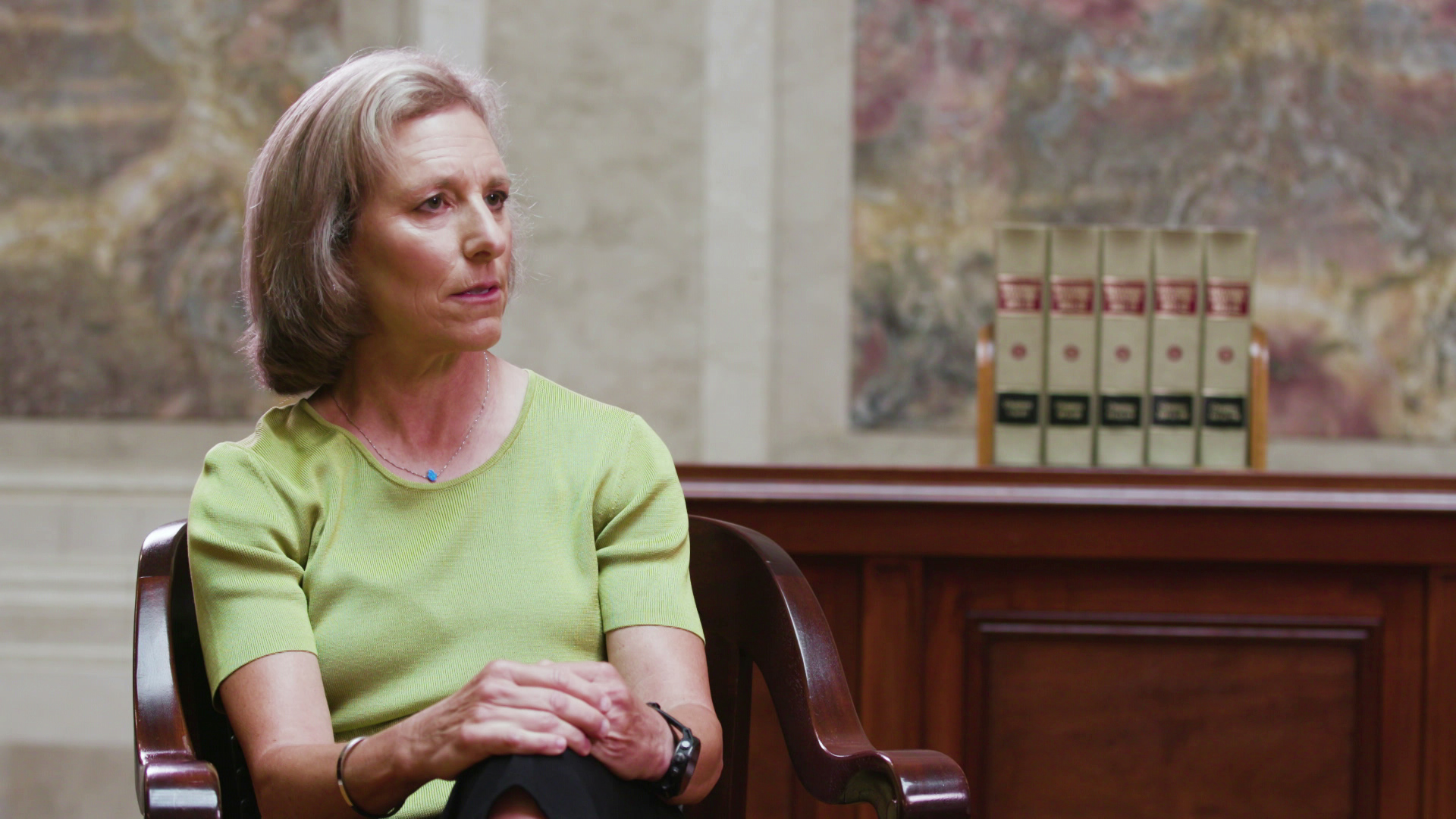 Jill Karofsky speaks while sitting in a room with a wood cabinet and a row of law books on its surface in the background.