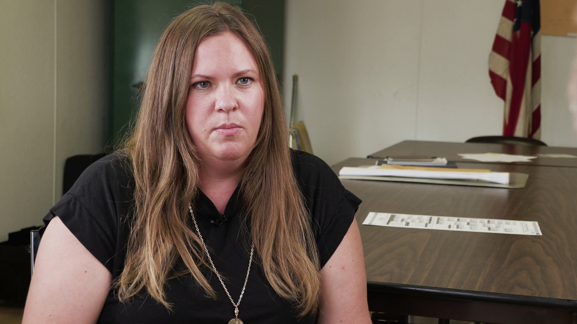 Melissa Kono speaks while sitting indoors with papers on a table and a U.S. flag in the background.