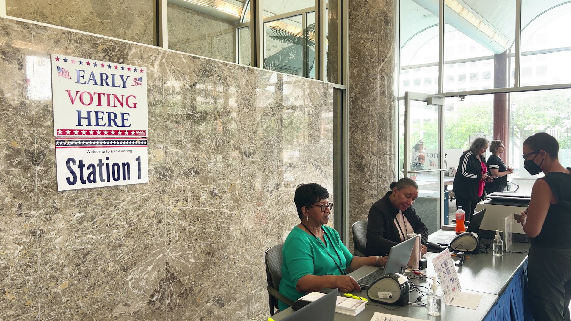 A person stands at the front of a table and faces two people seated on the other side, one using a laptop computer and the other writing in a room with large plate glass windows and marble masonry, with signs on one wall reading "Early Voting Here" and "Station 1."