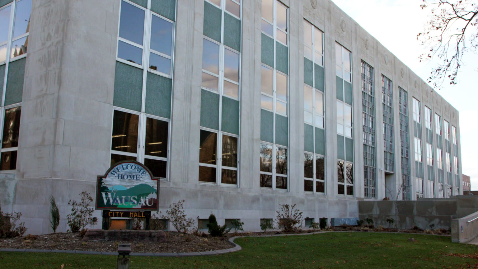 A weathered wood sign with stylized images of hills and trees and the words Welcome Home to Wausau and City Hall stands in a mulched garden bed in front of a multi-story masonry building with large glass windows in front of small grass lawn, a concrete ramp and tree branches.