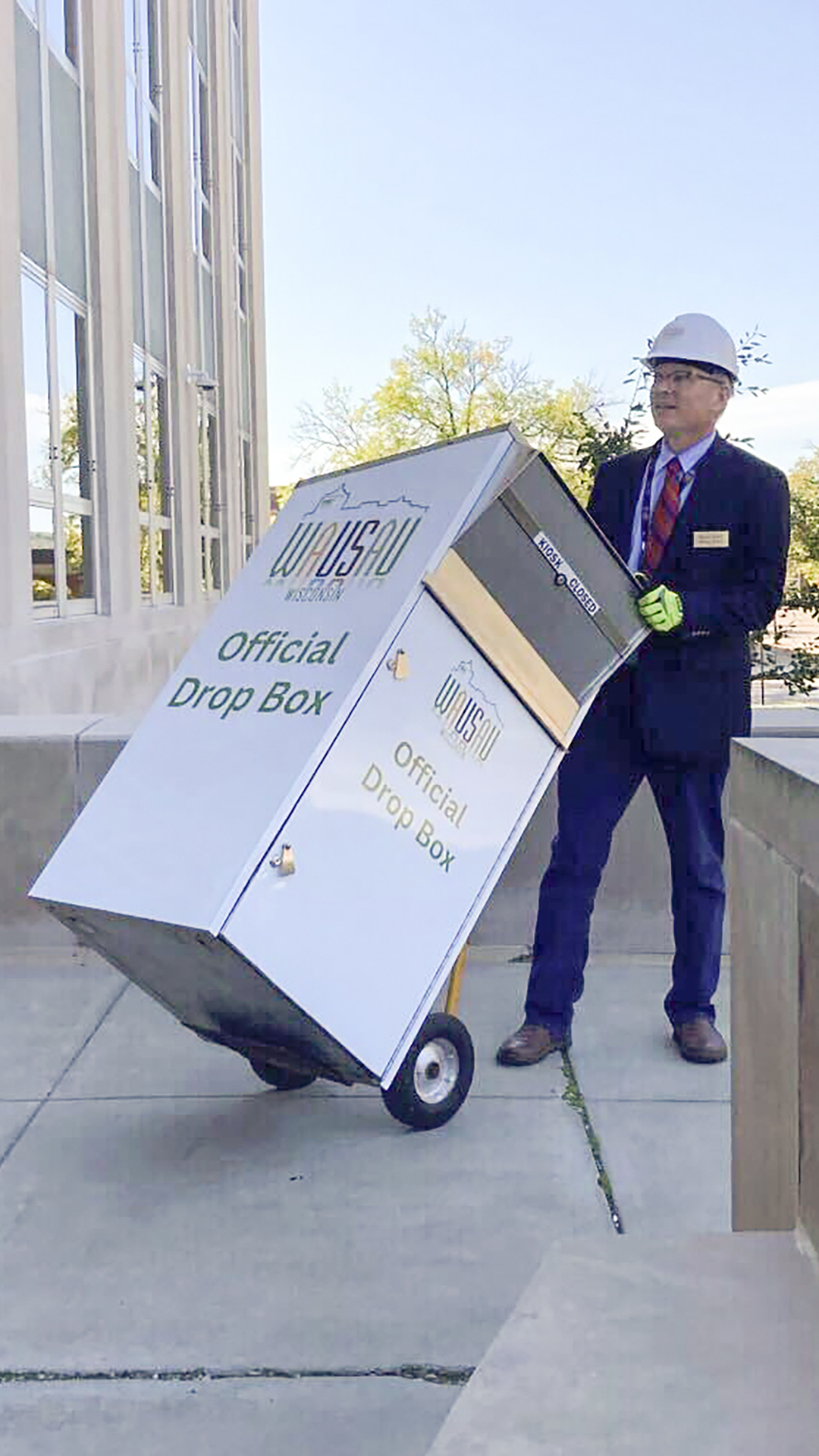 Doug Diny uses a two-wheeled dolly to move a metal box with a Wausau wordmark and the words "Official Drop Box" visible on two sides and a hinged door at its top across a concrete-paved patio of a multi-story masonry building with tall glass windows, with trees in the background.