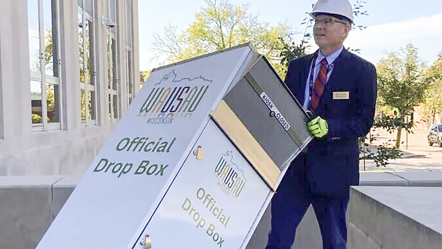 Doug Diny uses a two-wheeled dolly to move a metal box with a Wausau wordmark and the words Official Drop Box visible on two sides and a hinged door at its top across a concrete-paved patio of a multi-story masonry building with tall glass windows, with trees in the background.