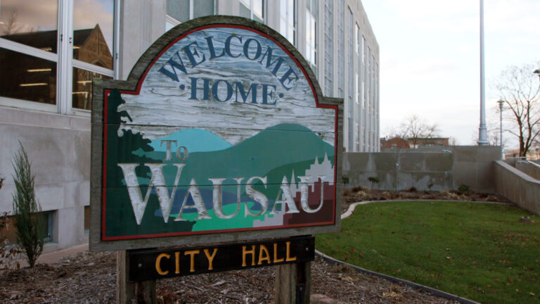 A weathered wood sign with stylized images of hills and trees and the words Welcome Home to Wausau and City Hall stands in a mulched garden bed in front of a multi-story masonry building with large glass windows, with a small grass lawn, a concrete ramp, flagpole and trees in the background.