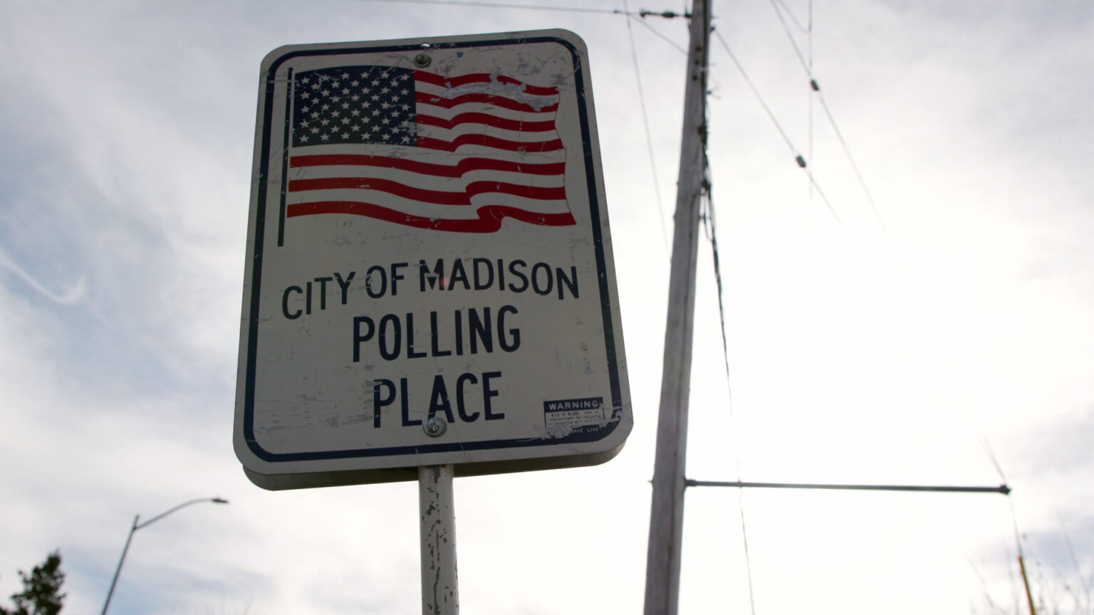 A metal sign with an illustration of a U.S. flag and the words City of Madison Polling Place is displayed on a metal pole, with a utility pole in the background under a sunlit cloudy sky.