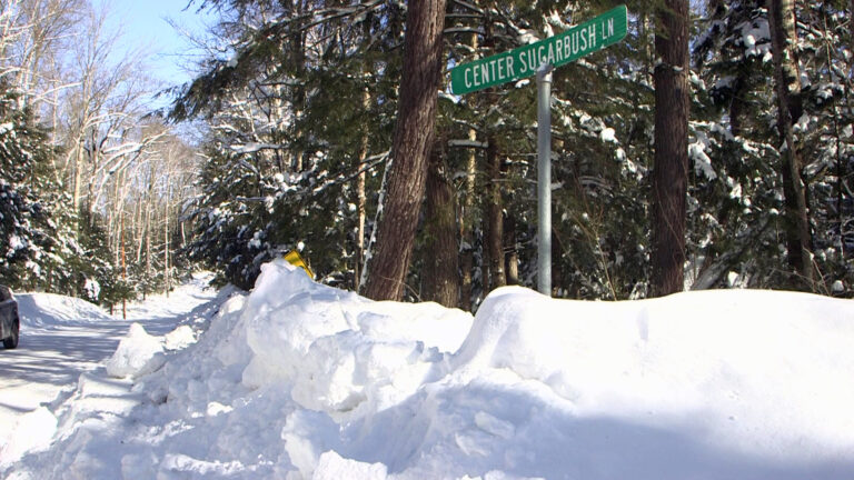 A street sign that reads Center Sugarbush Ln stands on a metal pole in the middle of a snowbank on the side of a snow-packed road, with snow-covered conifer trees in the background.