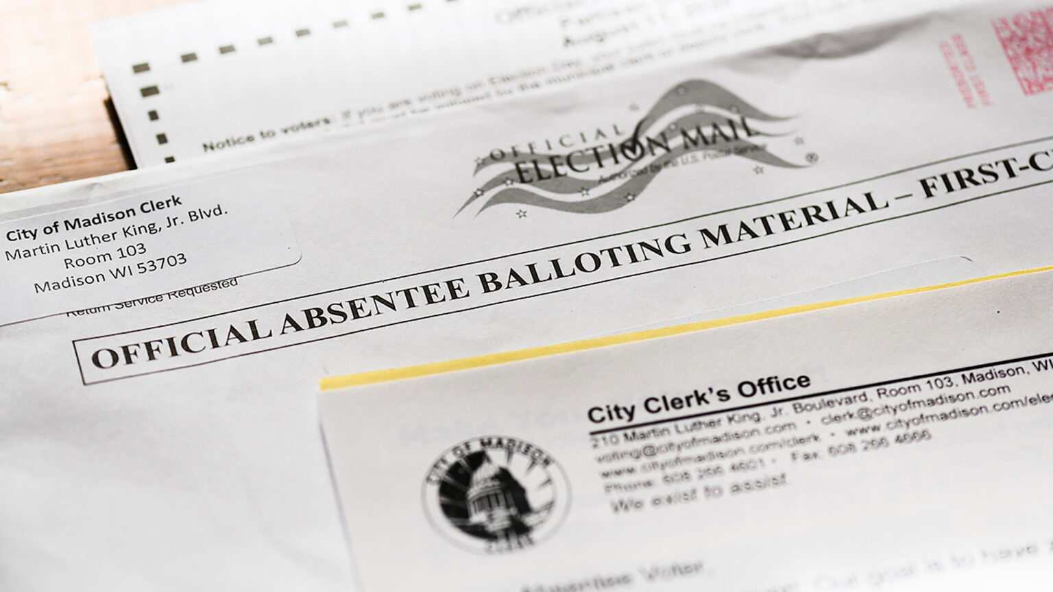 An envelope with words reading Official Absentee Balloting Material and a return address sticker from the City of Madison Clerk sits on a wood surface between a letter from the City's Clerk's office and a ballot.