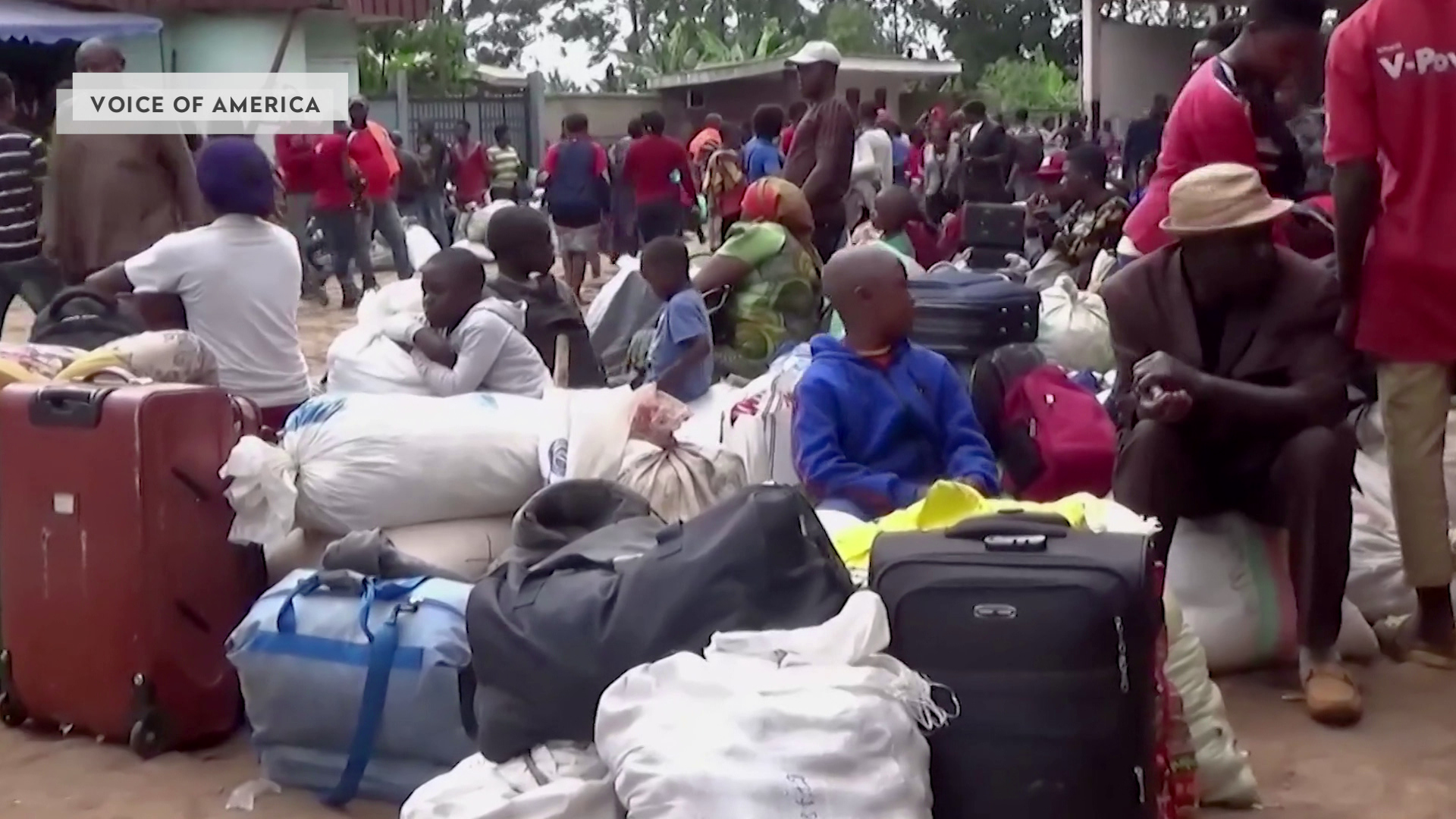 Numerous people sit and stand among piles of suitcases and bags outside with tress in the background.