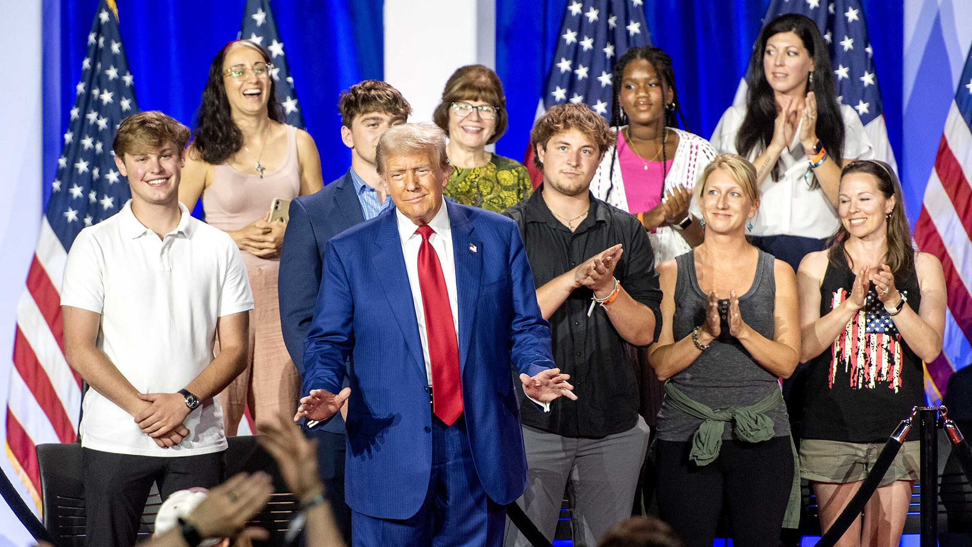 Donald Trump gestures with both hands facing palm down in front of him while nine other people standing behind him applaud, in a room with U.S. flags in the background.