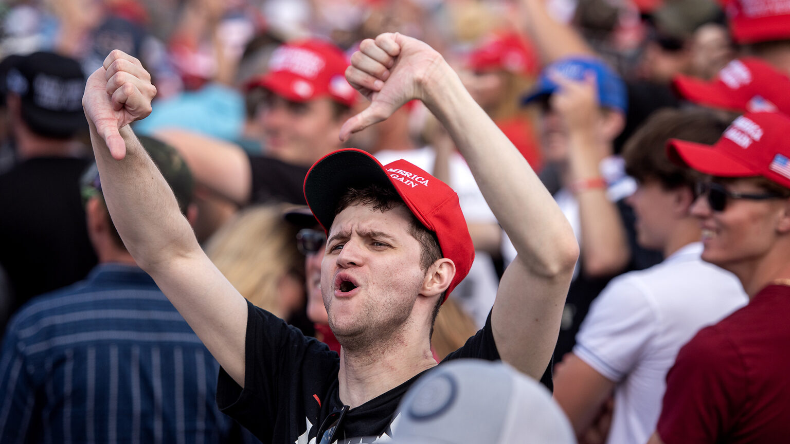 A man wearing a Make America Great Again cap makes thumbs down gestures with both hands raised above his head while shouting, with other people standing in the background.