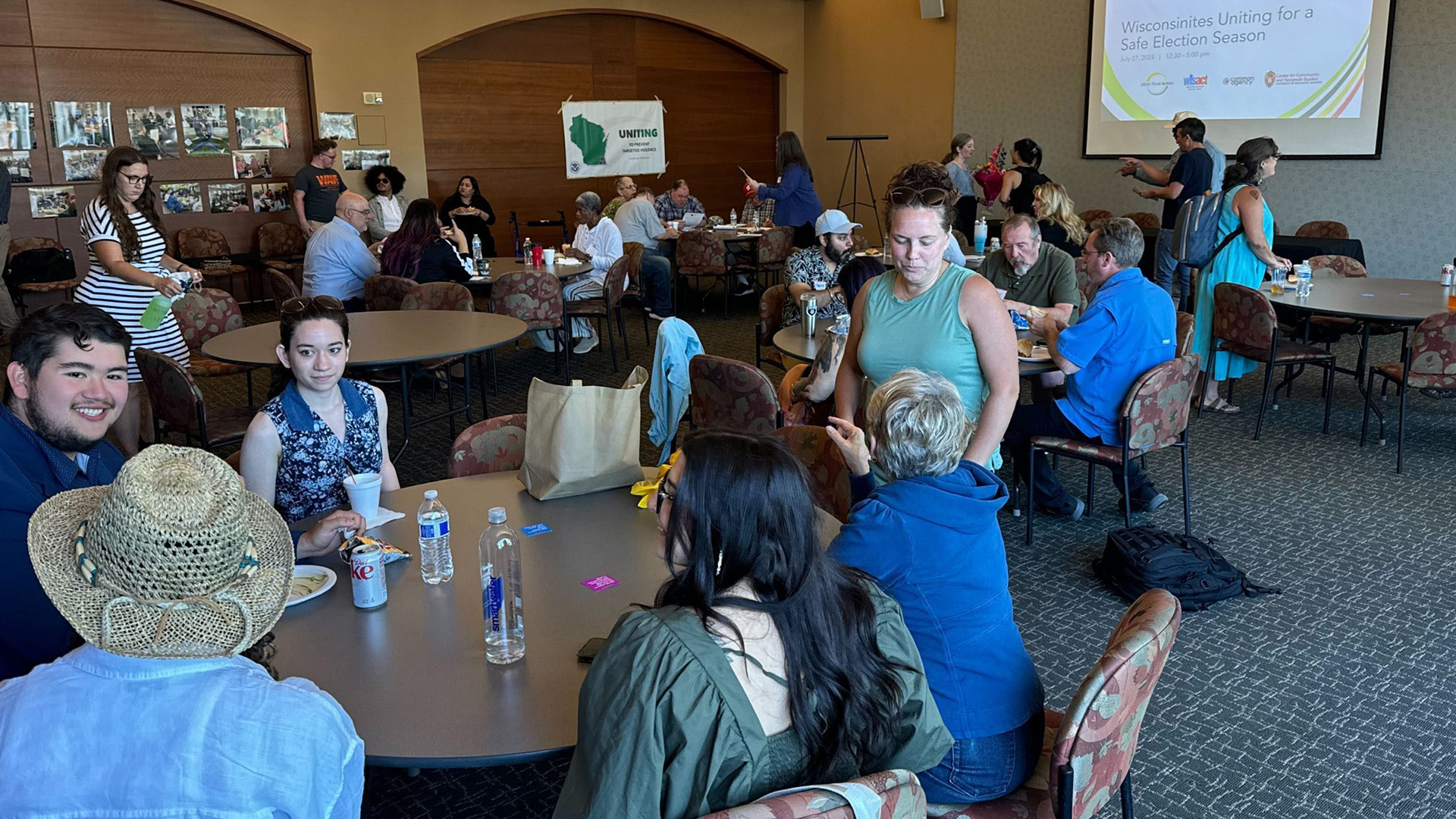 People sit in chairs and stand around round tables in a room with a patterned carpet, wood-paneled and wallpapered walls, and a projection screen showing a slide with the words "Wisconsinites Uniting for a Safe Election Season"