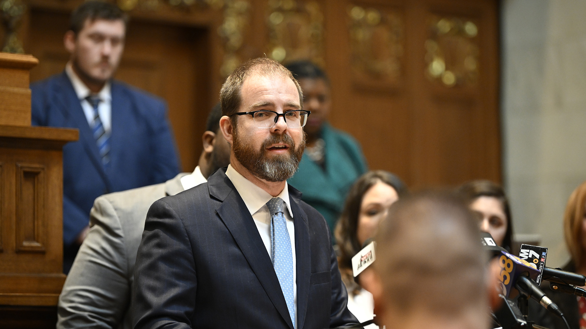 Ryan Clancy speaks into multiple microphones with the flags of different media organizations while standing in front of and facing multiple people, in a room with a wood legislative dais in the background.