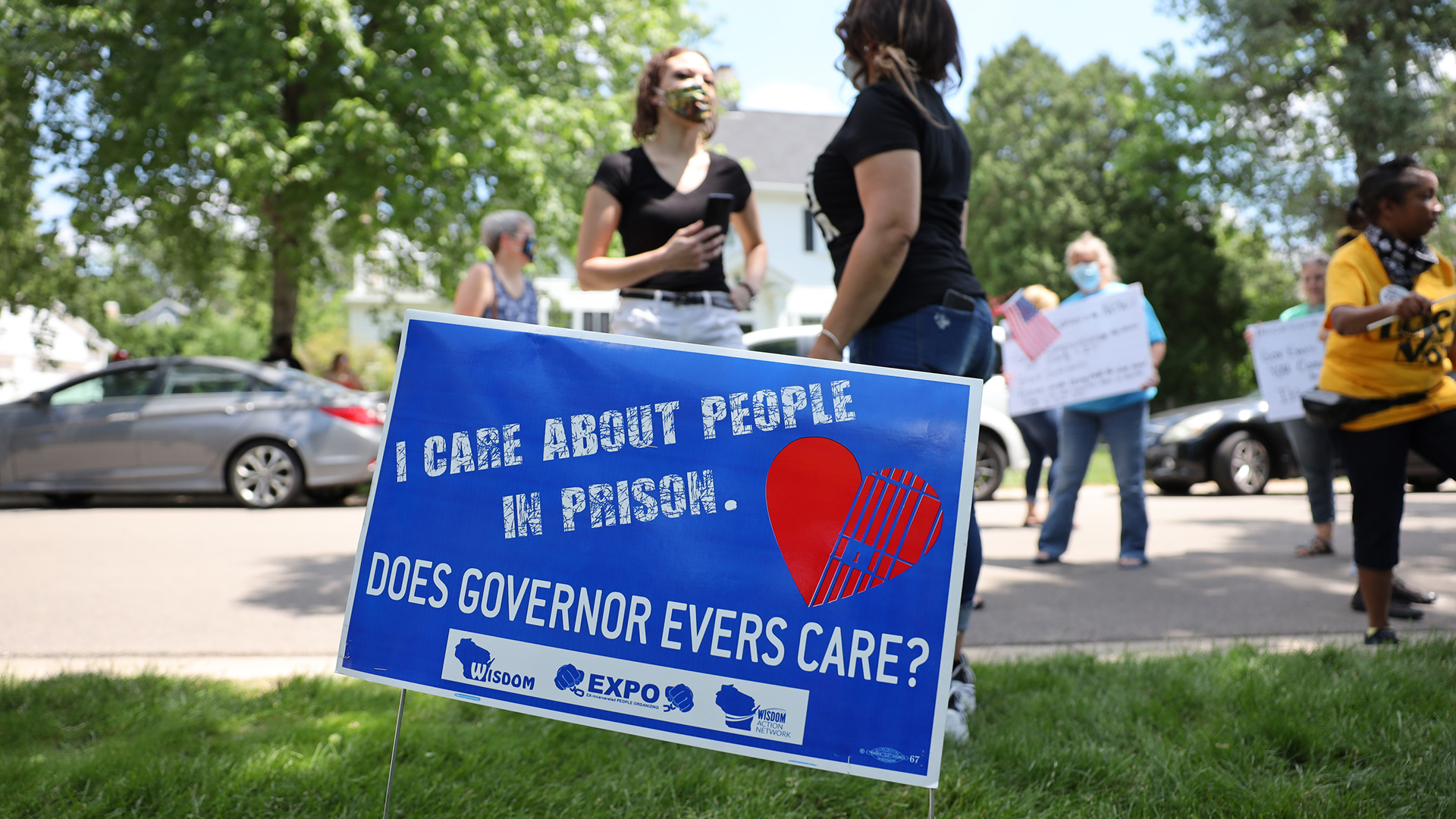 A cardstock political yard sign with the words "I Care About People in Prison." and "Does Governor Evers Care?" is placed in a lawn in front of a road with multiple people standing in it in front of parked vehicles, with trees and a building in the background.