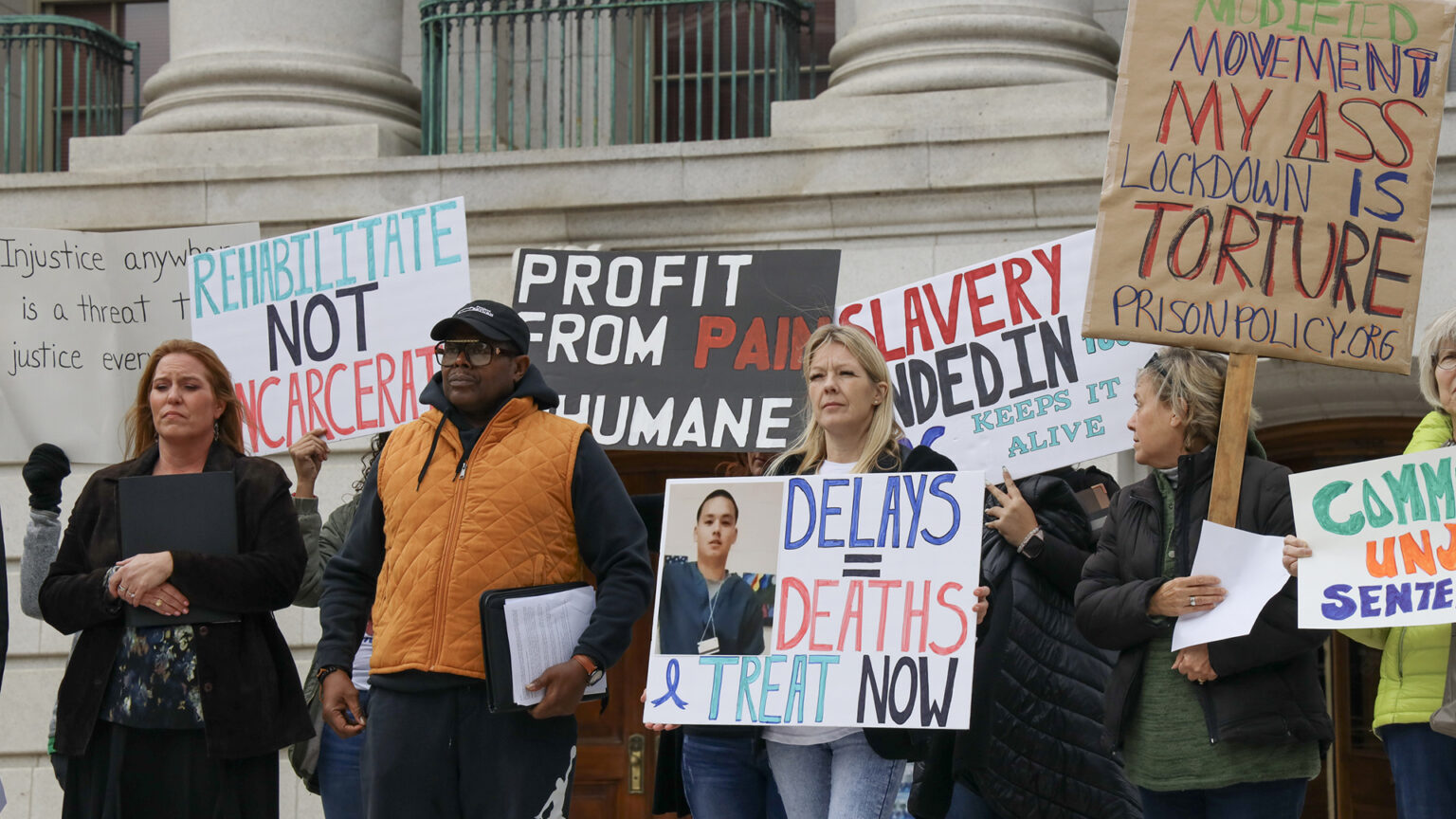 Multiple people stand holding hand-drawn signs, including ones reading Rehabilitate Not Incarcerate, Delays = Deaths Teat Now, while standing in front of a marble masonry building beneath the base of multiple columns with copper patina metal fences between them.