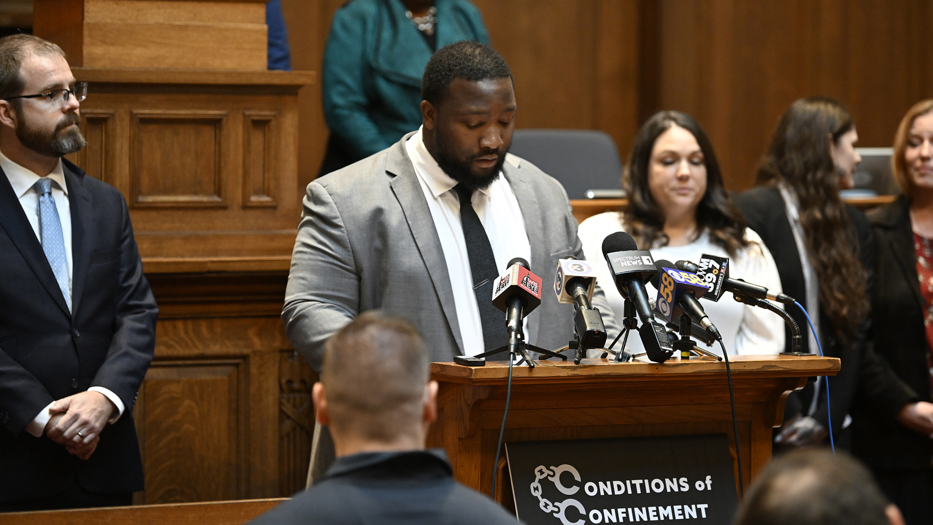 Darrin Madison speaks into multiple microphones with the flags of different media organizations attached to a wood podium with a sign on its front with the words "Conditions of Confinement" with the two letter Cs illustrated as handcuffs, while standing in front of and facing multiple people, in a room with a wood legislative dais in the background.