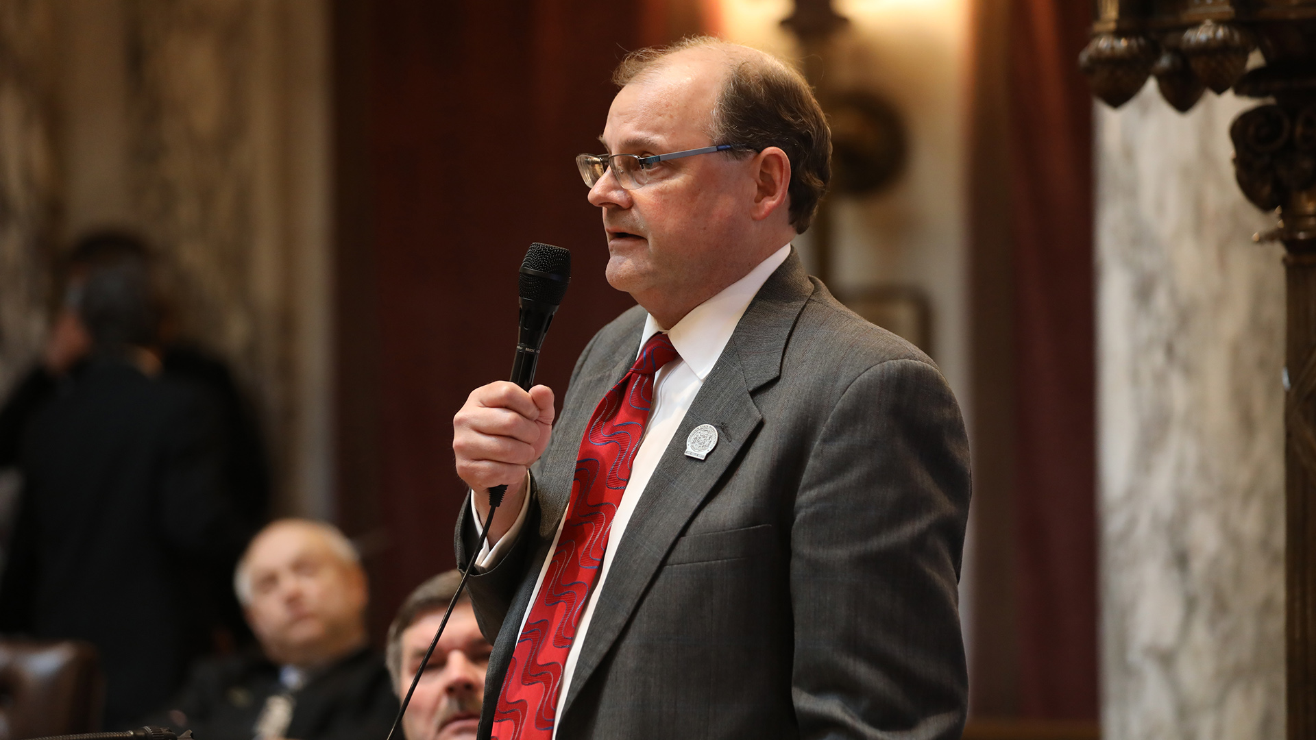 Michael Schraa holds and speaks into a wired microphone while standing in front of seated people in a room with marble masonry, wood wall panels and brass light fixtures.