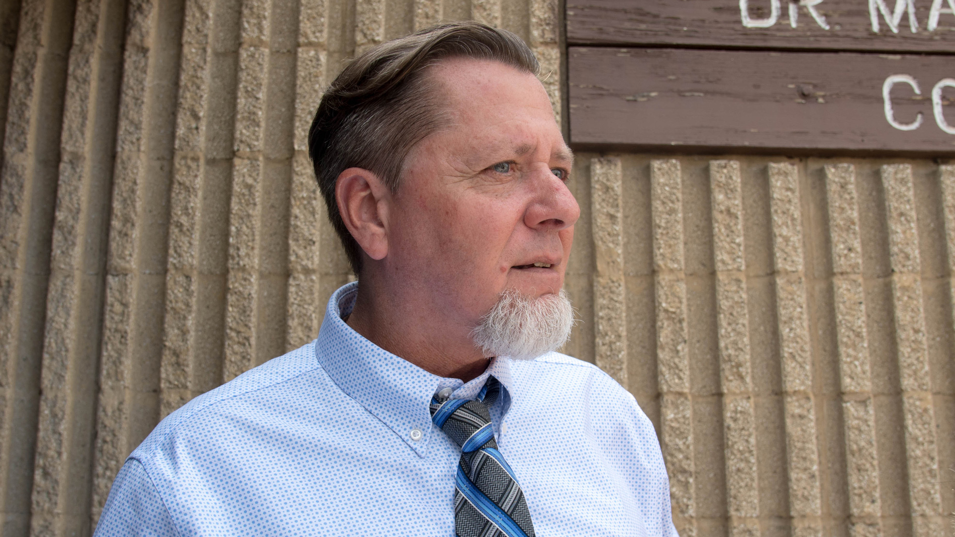 Lonnie Story stands outside in front of a masonry wall with a wood sign attached to its surface.