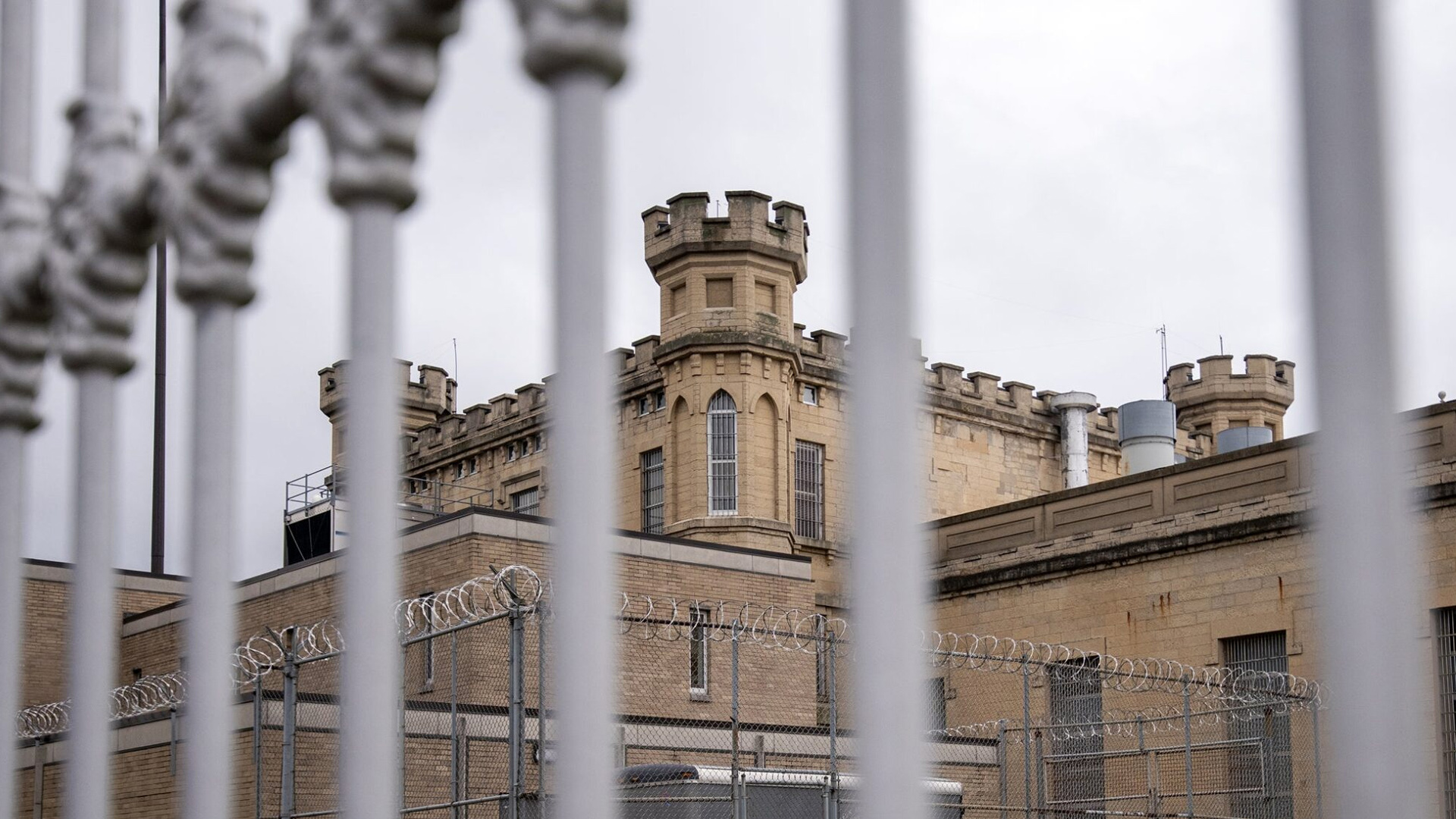 Multiple brick and masonry buildings, including a central structure topped by crenelated towers, stand next to an outdoor area ringed with a chain-link fence topped by razor wire, as seen through out-of-focus vertical bars.