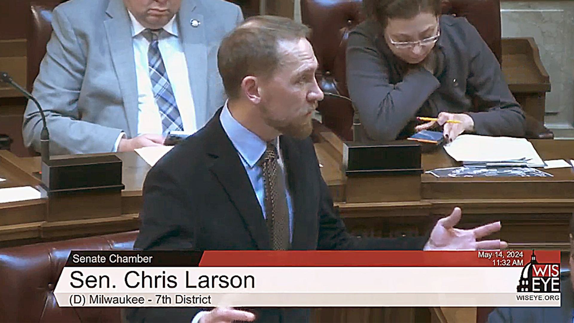 A video still image shows Chris Larson standing and speaking with other people seated along a curved row of desks in the background, with a video graphic at bottom including the text "Sen. Chris Larson" and "(D) Milwaukee - 7th District."