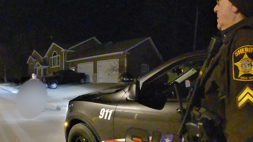 A Lafayette County Sheriff's Office officer stands in front of a squad SUV parked on a road with its headlights illuminating an area of pavement that is blurred out with only the head of a standing individual visible, with a house and truck parked in its driveway in the background.