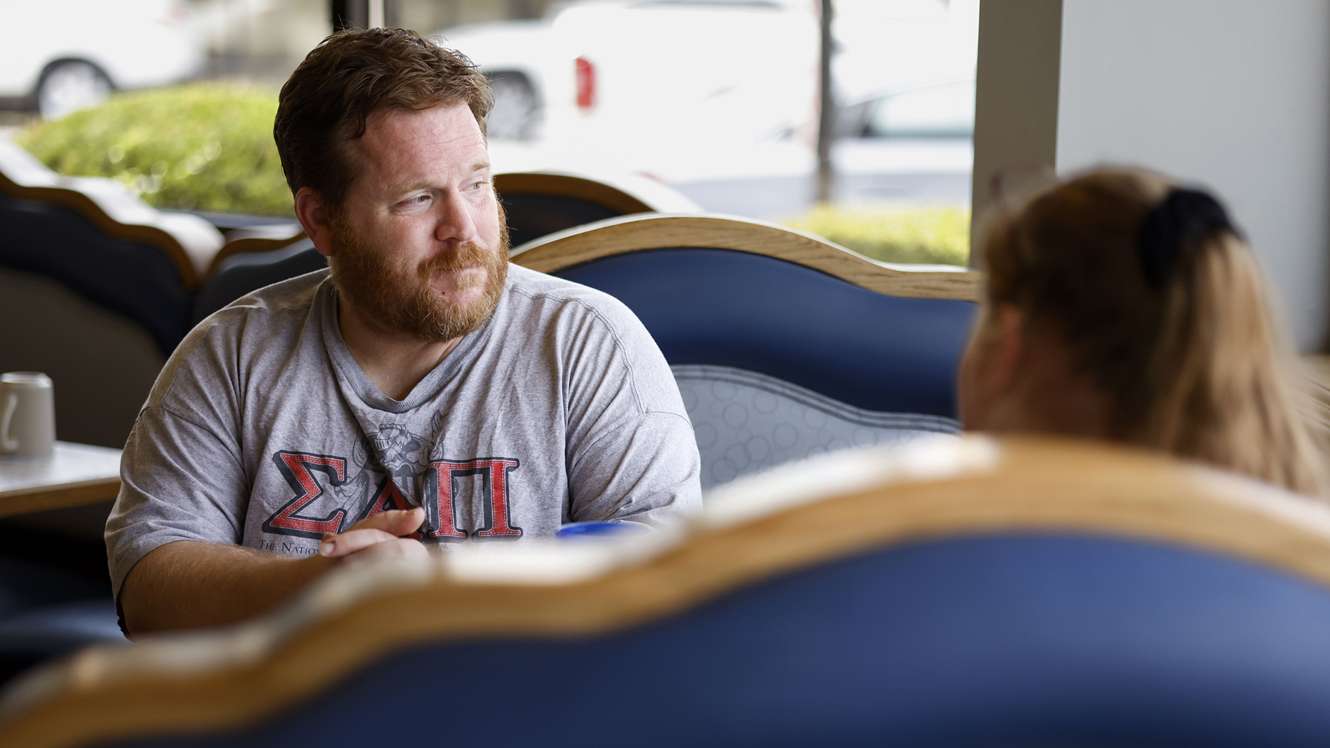 Joseph Crowley sits in a wood and vinyl restaurant booth and faces another person, with out-of-focus parked vehicles visible through plate-glass windows in the background.