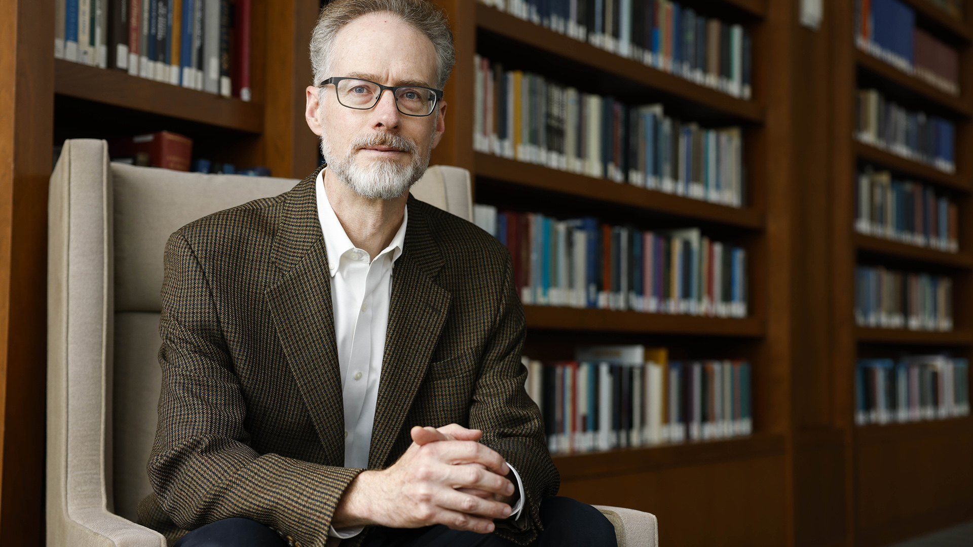 Michael O'Hear poses for a portrait while seated in an armchair with wood bookcases in the background.