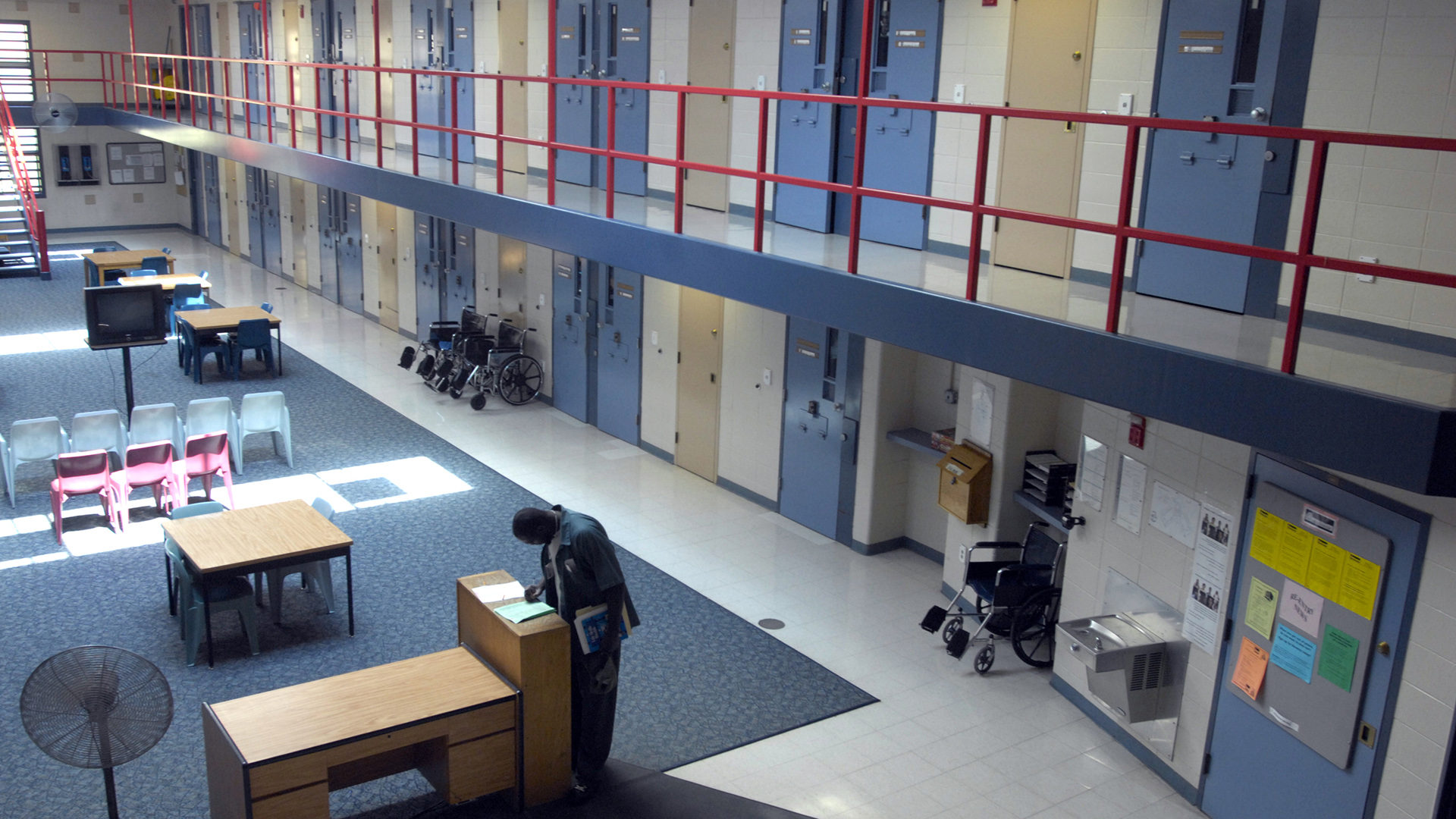 A persons stands and looks at a paper on top of a wood shelf next to a desk in an interior courtyard space with more desks and chairs, a wall-mounted drinking fountain, and three wheelchairs next to a wall with multiple closed metal doors with small vertical windows, below a second level with walkway with a metal railing providing access to another row of closed metal doors.