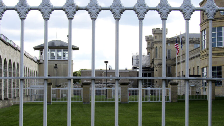 Multiple brick and masonry buildings, including a structure topped by crenelated towers and a courtyard with a mown lawn, as seen through vertical bars.