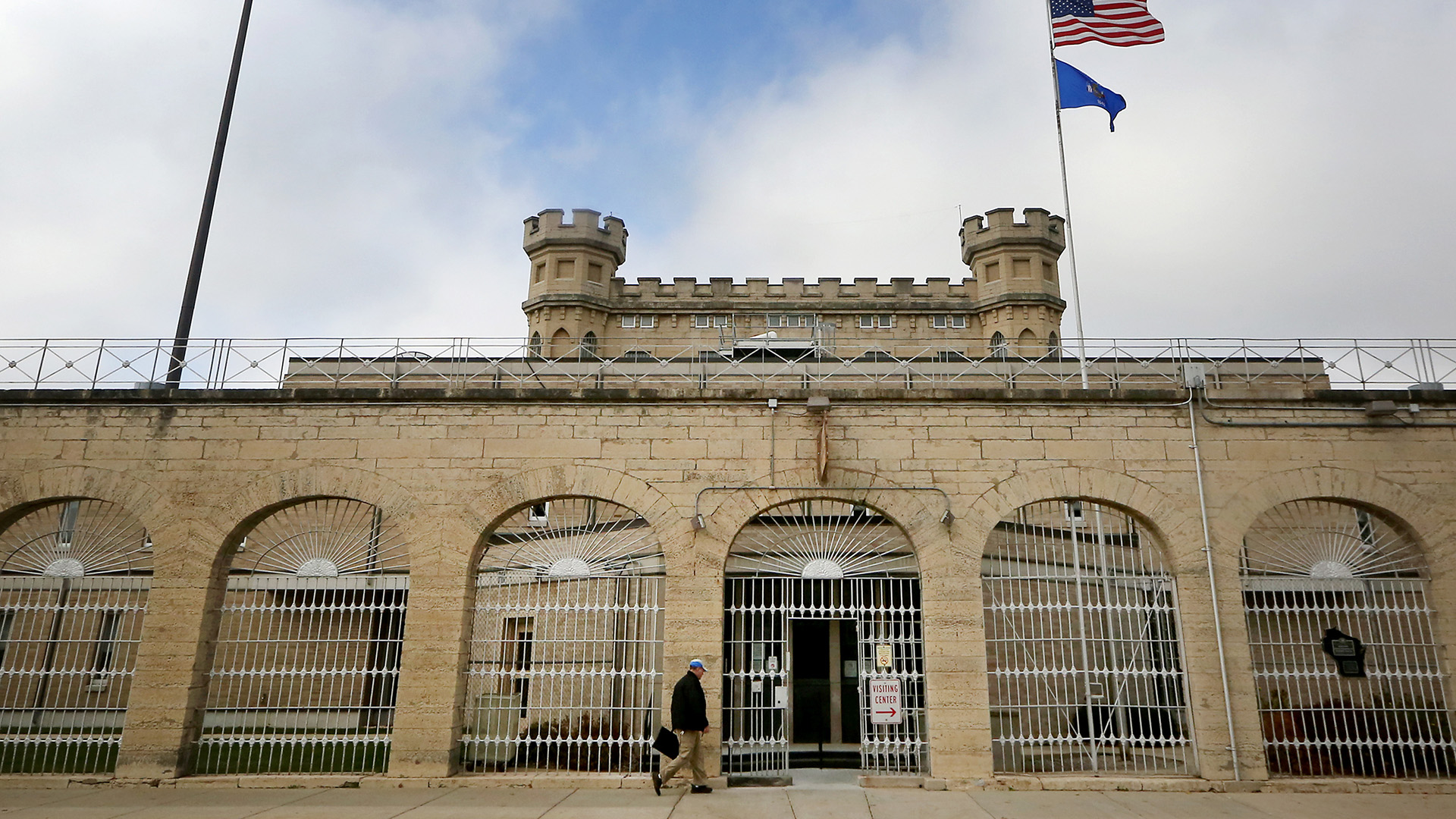 A person walks along a sidewalk toward gate in a barred fence that stands open in a central archway of a masonry wall standing outside additional brick and masonry buildings, with the U.S. and Wisconsin flags on a flagpole standing in front of a central structure topped by two crenelated towers, under an mostly cloudy sky.