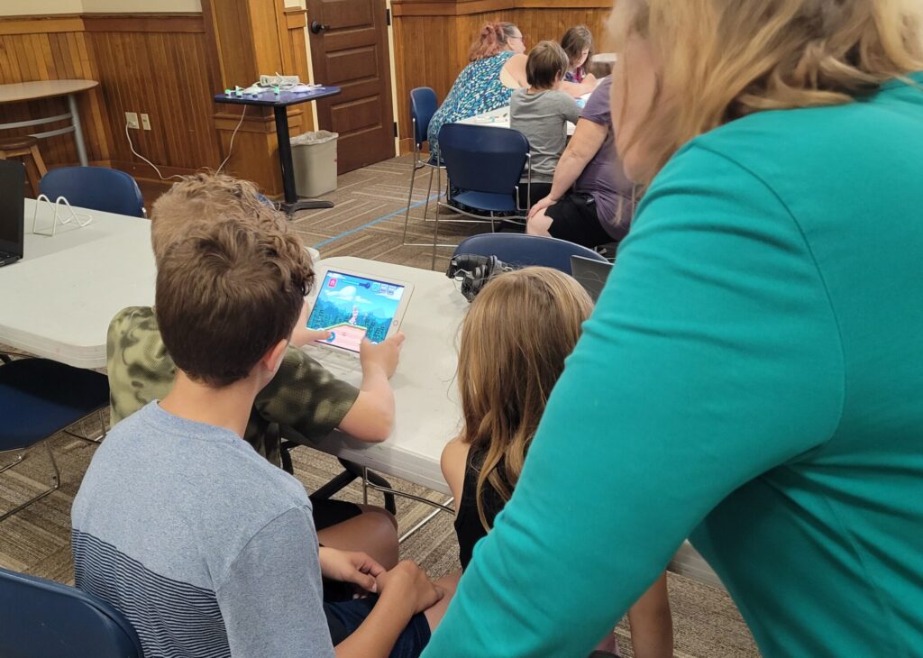 Three children sit at a table playing an educational game on a tablet. A grown-up looks over their shoulders.