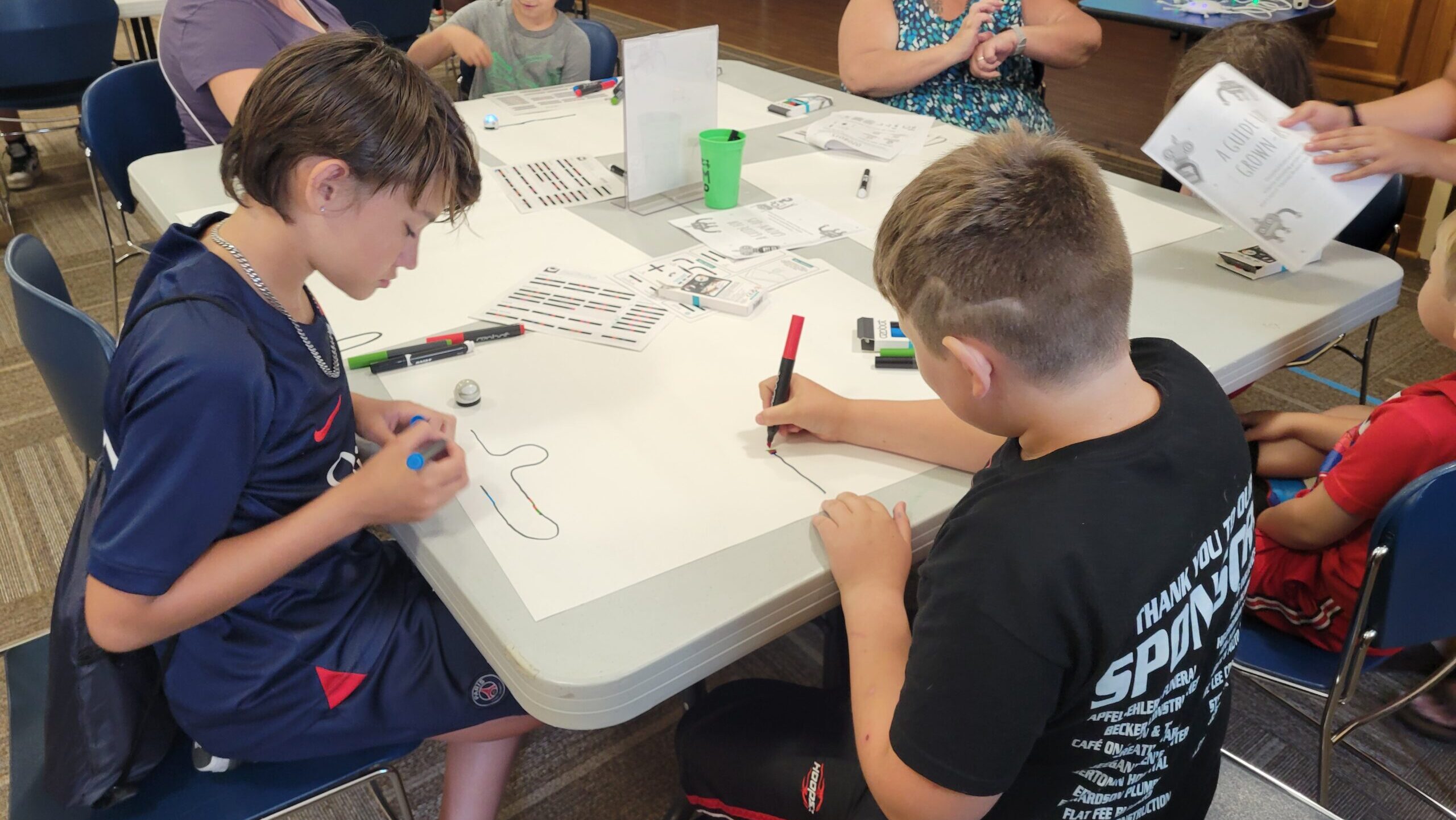 Kids and grown-ups sit at a large table drawing on paper spread across the table.