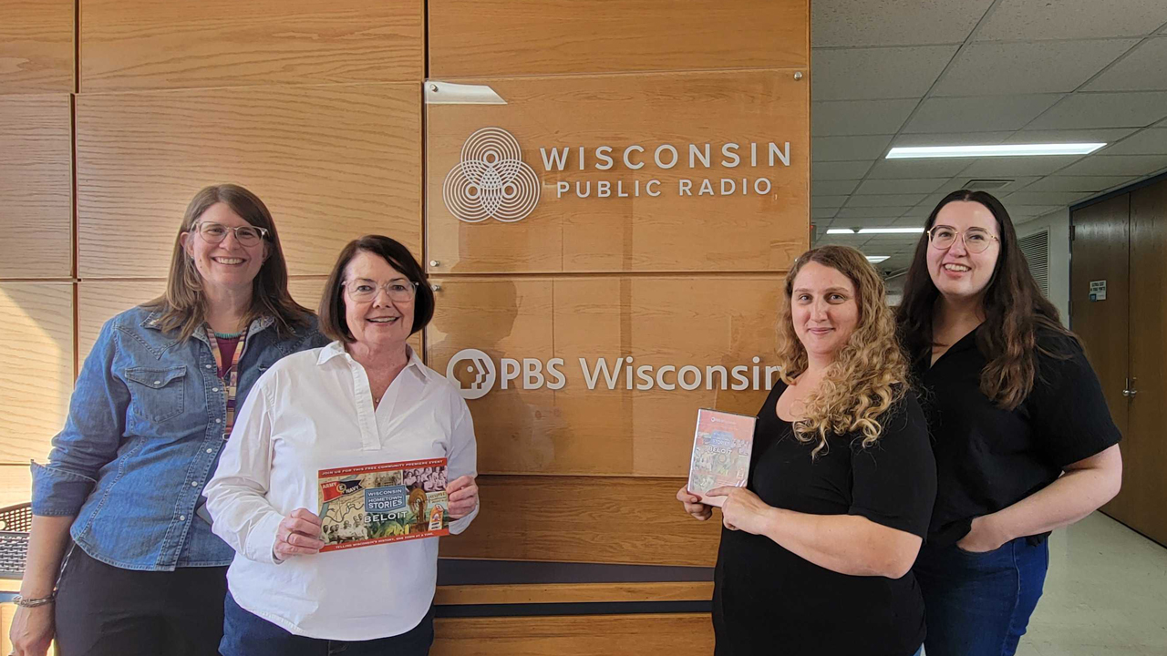 Four women smile, as they pose standing in front of the Wisconsin Public Radio and PBS Wisconsin sign of the Wisconsin Public Media lobby.