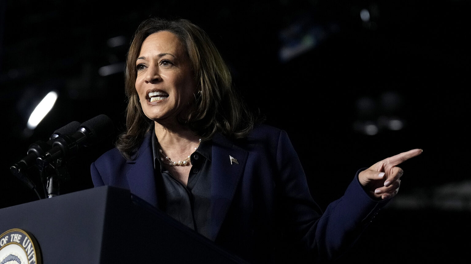 Kamala Harris points to the side with her left index finger while standing behind a podium and speaking into two mounted microphones.