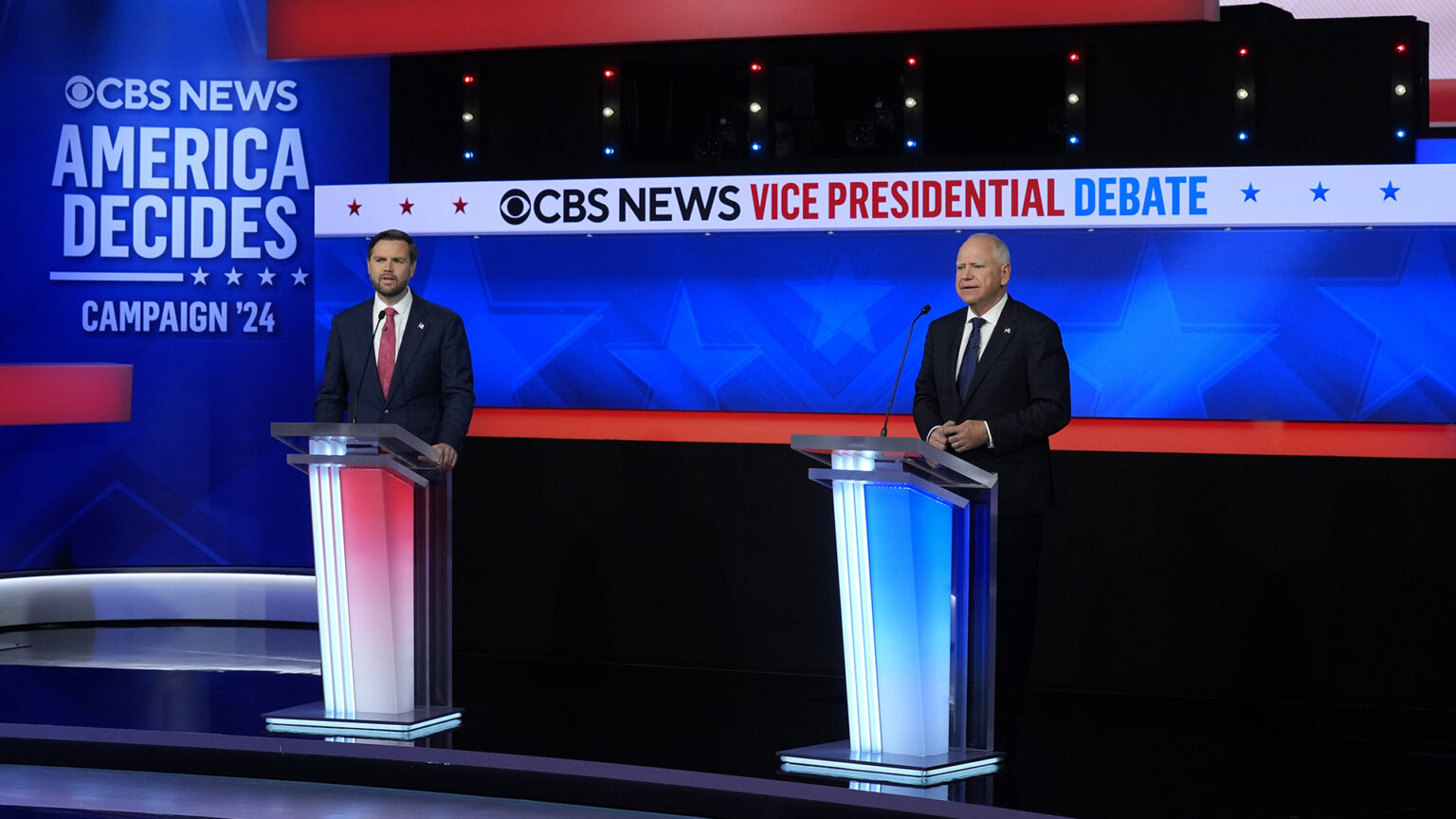 JD Vance and Tim Walz stand at two podiums in a television studio with illuminated signs reading CBS News Vice Presidential Debate and CBS News America Decides Campaign '24.