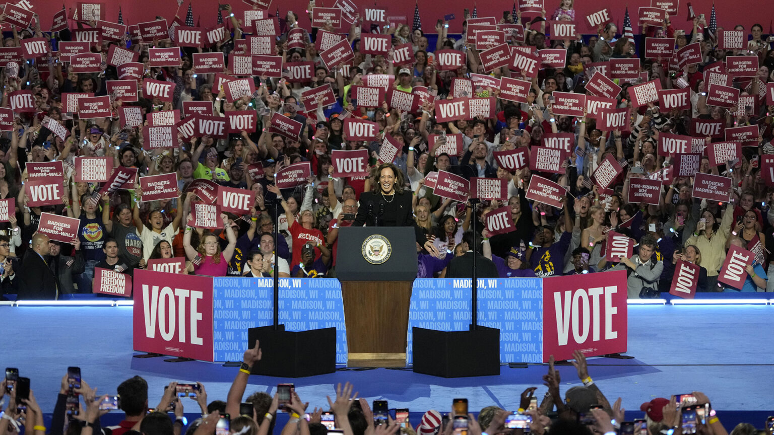 Kamala Harris speaks while standing behind a podium with the Seal of the Vice President of the United States affixed to its front, with two teleprompters and mirrors on stands on either side, and a low backdrop with signs reading Vote and Madison, WI behind her on a stage, with cheering audience members holding signs reading Vote, Badgers for Harris-Walz and Freedom Freedom Freedom on bleachers in the background, and cheering audience members standing below the stage facing her and recording photos and videos with smart phones in the foreground.