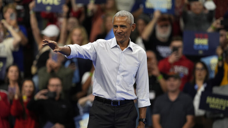 Barack Obama waves with his right hand while standing in a room with out-of-focus people standing, holding signs, taking photos and cheering in the background.