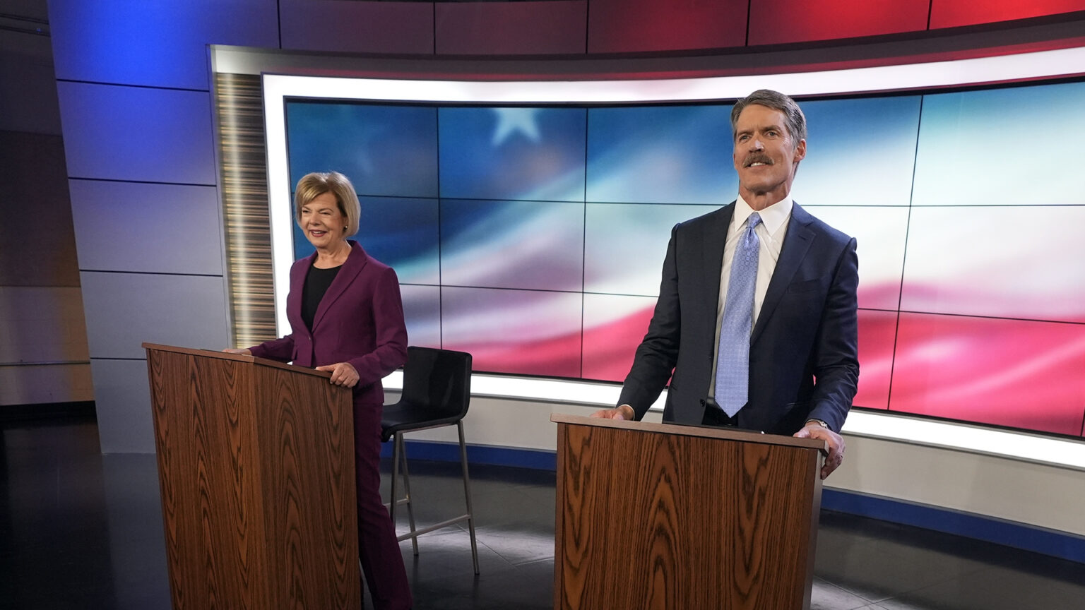 Tammy Baldwin and Eric Hovde stand at and rest both of their hands on the top of two wood podiums, in a room with a bank of monitors behind them showing a stylized U.S. flag and overhead blue and red lighting.