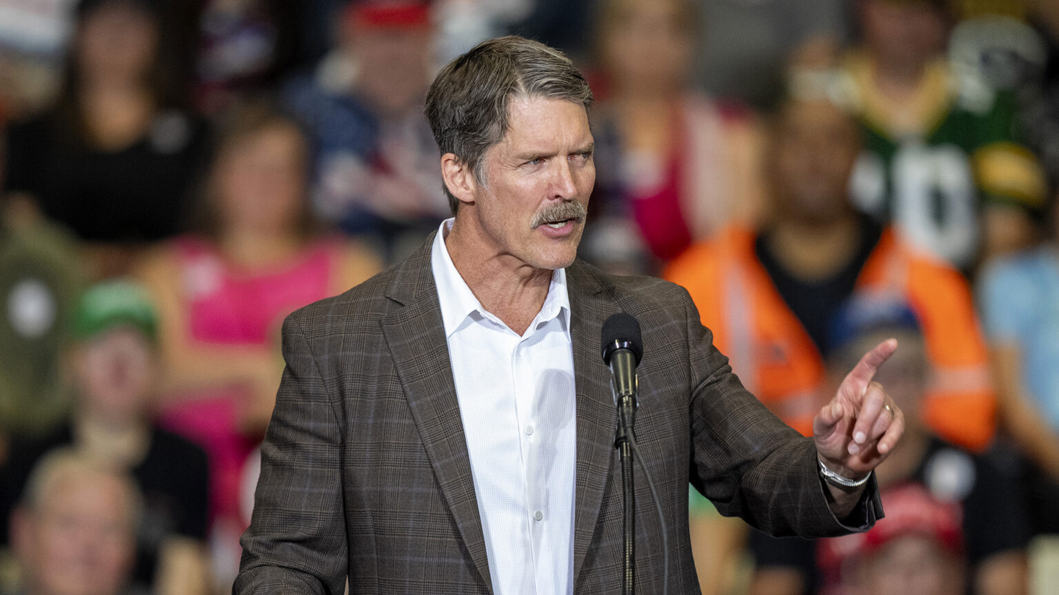 Eric Hovde gestures with his left hand while standing and speaking into a microphone mounted on a stand, with out-of-focus people standing behind him.