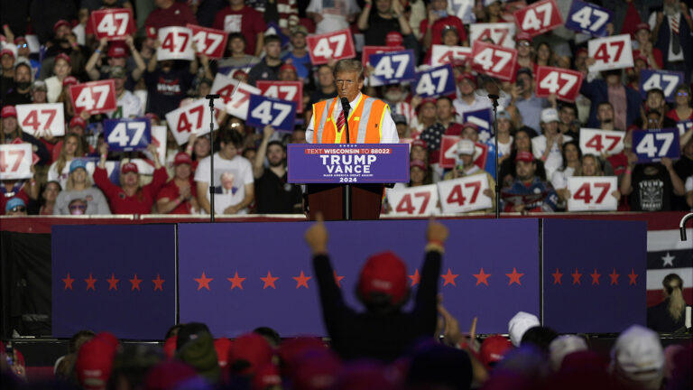 Donald Trump speaks into a microphone while standing behind a podium with a sign with the words Trump Vance 2024 and a text message number affixed to its front, with two teleprompter mirrors on stands on either side, and a low backdrop in front of him on a stage, with cheering audience members holding signs reading 47 on bleachers in the background, and cheering audience members standing below the stage facing him in the foreground.