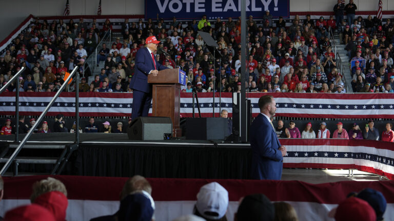 Donald Trump speaks into microphones mounted to a wood podium while standing on a temporary stage while facing multiple stage floor monitors and two teleprompter mirrors and the frame of protective shielding, with one person standing at the corner of the stage, people standing behind red, white and blue bunting attached to modular metal fencing and other people seated on temporary bleachers with bunting on its bottom level and a sign at the top reading Vote Early!, and with out-of-focus people seated behind bunting and facing the stage in the foreground.