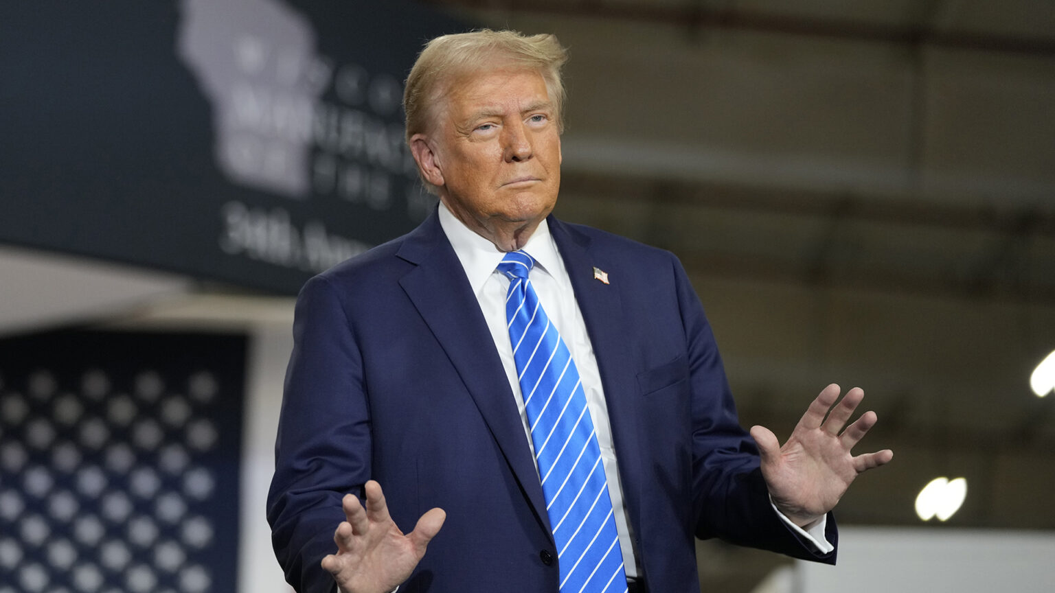 Donald Trump holds both hands in front of him while standing in a warehouse with the out-of-focus stars of a U.S. flag and a banner with an outline of Wisconsin in the background.