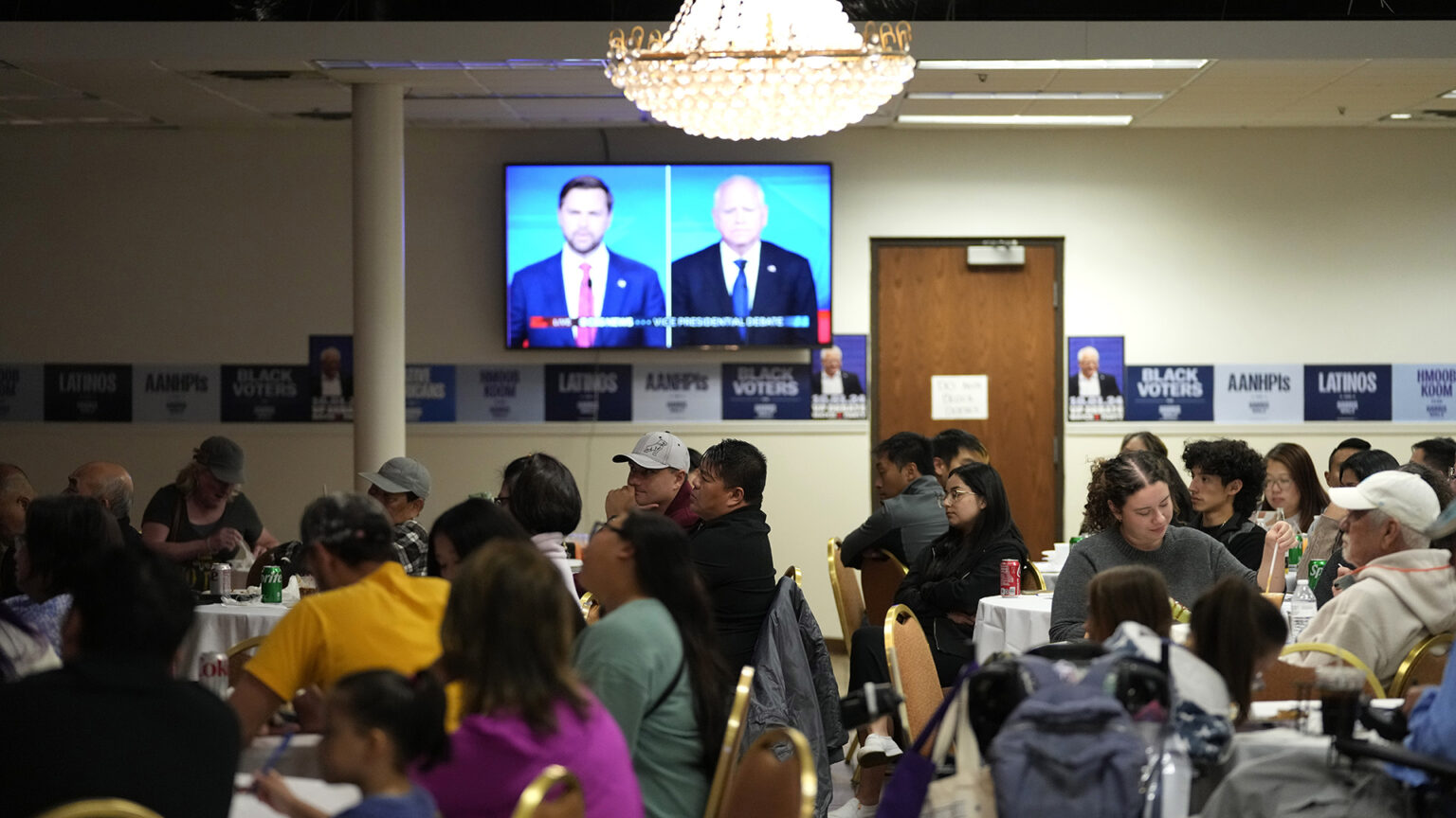 People of different ages sit in metal and upholstered banquet stack chairs at round tables below an illuminated glass chandelier in a room with fluorescent lights in a suspended ceiling, a closed wood door with a hydraulic closer, a row of paper political signs displayed on a rear wall and a television monitor showing JD Vance and Tim Walz in a split screen.